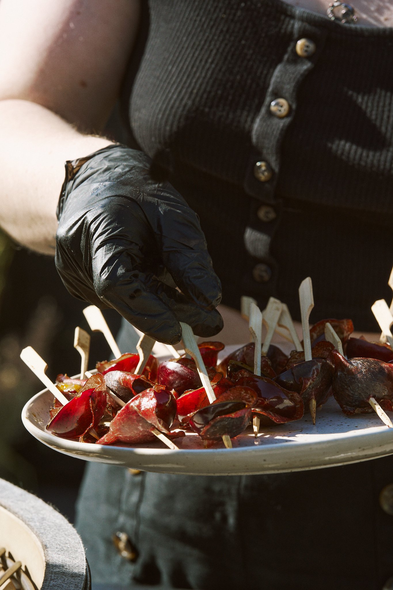 A person wearing a black sleeveless shirt and black gloves is holding a plate of roasted red peppers with skewers in them.