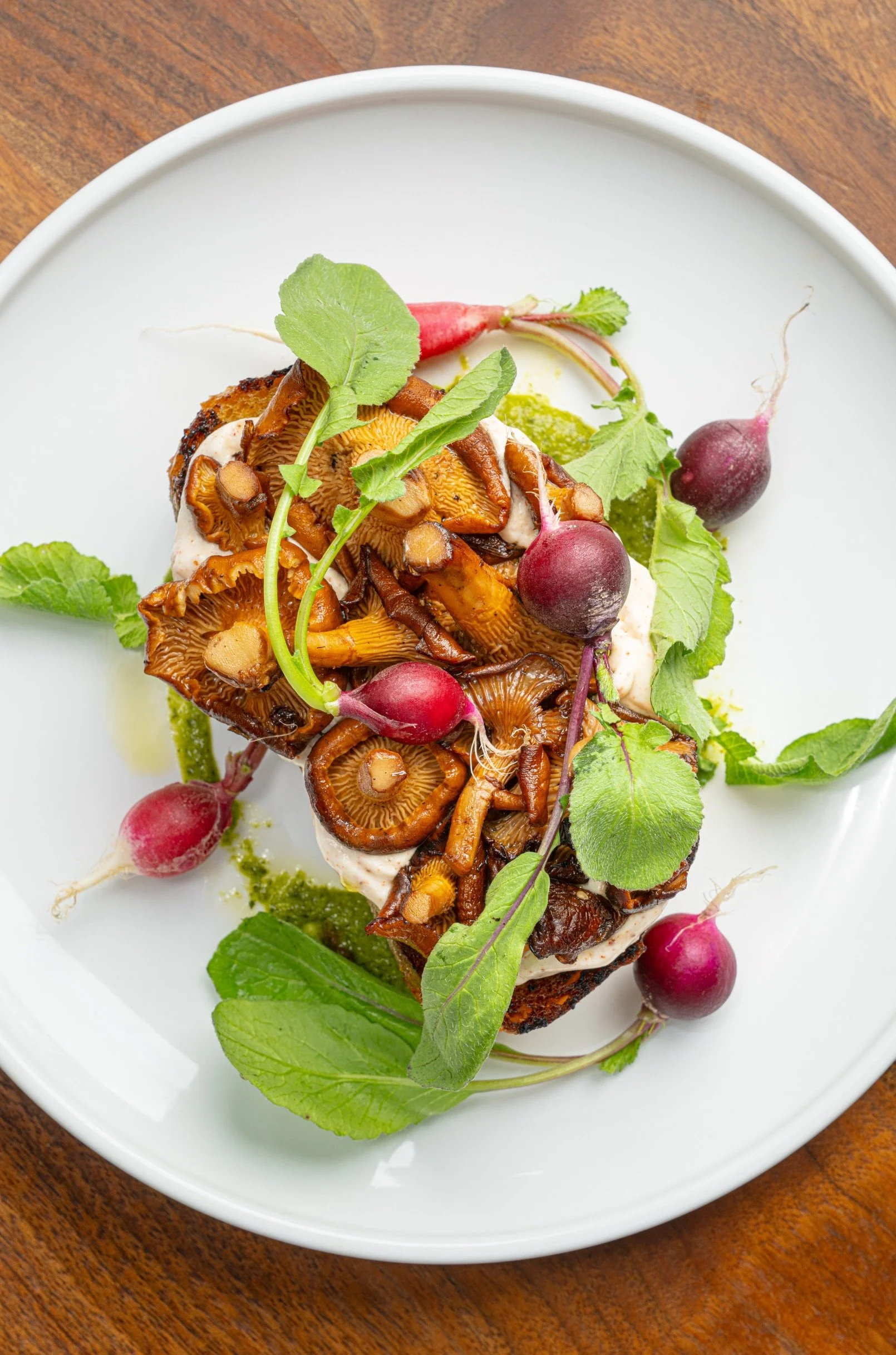 A white plate with cooked mushrooms, garnished with radishes, green leaves, and a green sauce.
