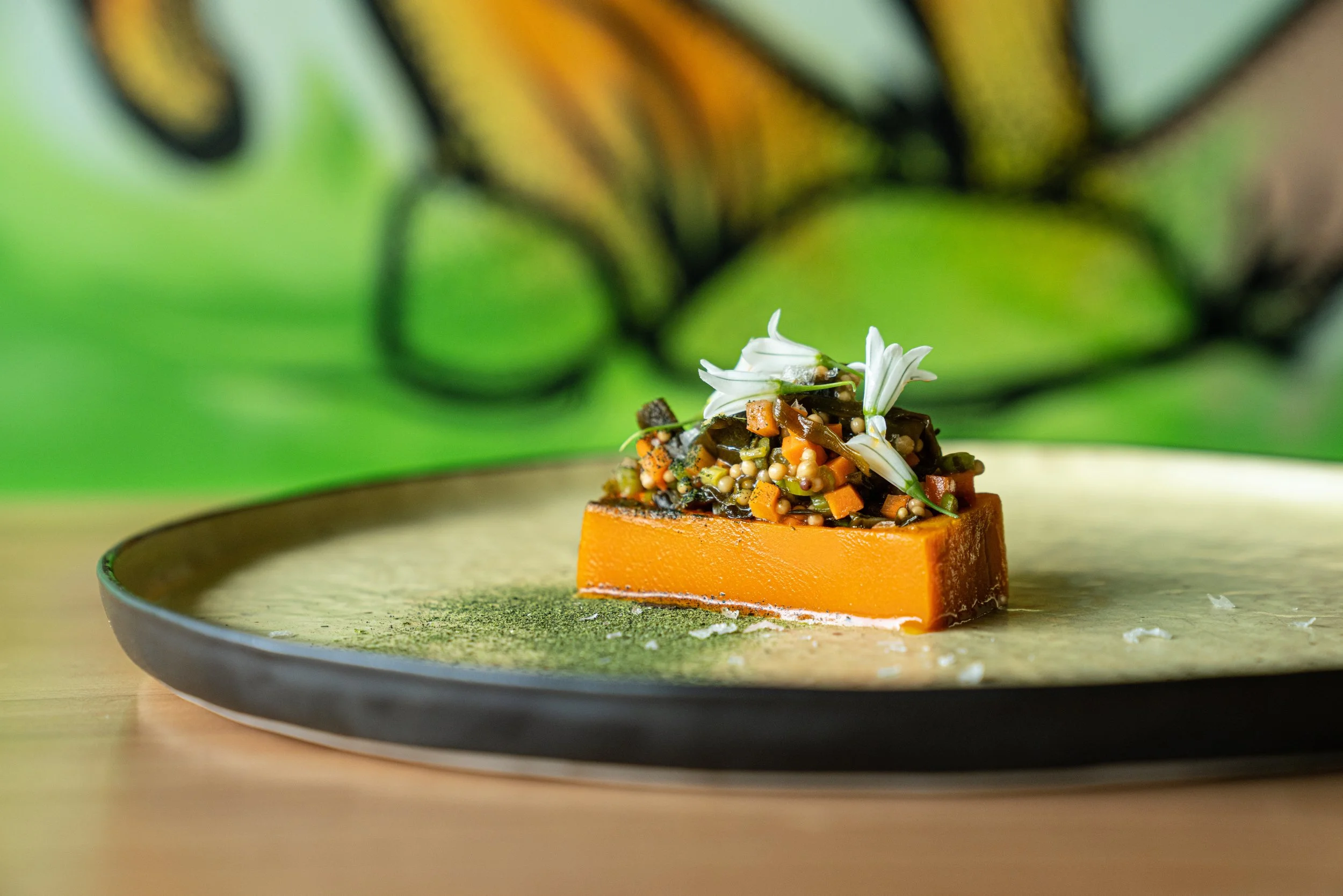 A plated dish features a rectangular piece of orange-colored food topped with a mixture of small diced vegetables, green herbs, and white edible flowers, presented on a round black and gold plate.