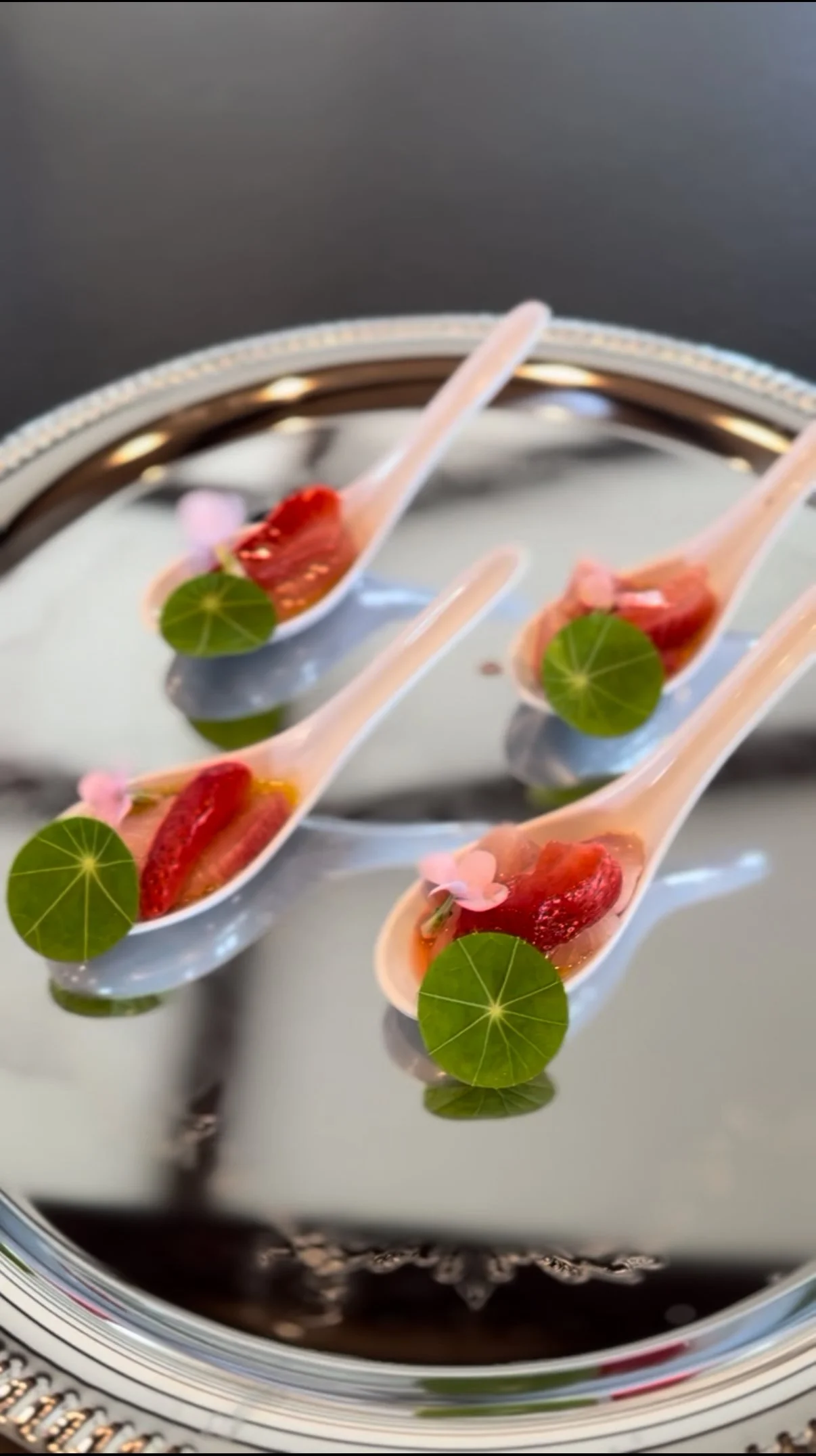 Three porcelain spoons filled with sliced strawberries, green leaves, and pink edible flowers, displayed on a reflective silver tray.