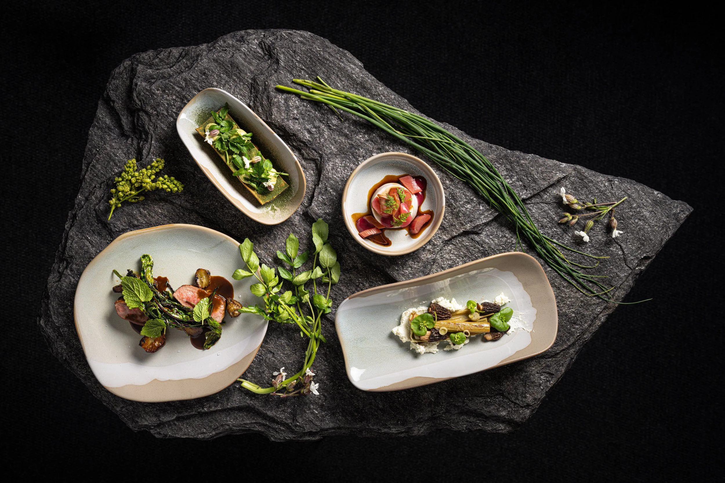 Assorted gourmet dishes with greens and herbs on decorative plates, set on a textured black stone slab with additional herbs and flowers for presentation.