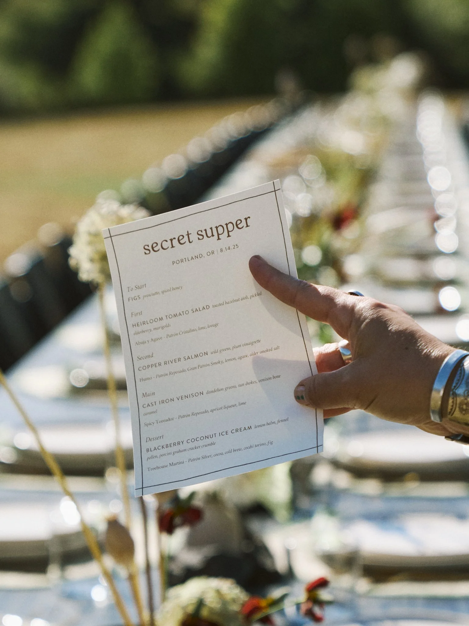 A person holding a menu titled 'Secret Supper' at an outdoor event, with a blurred picnic table setting in the background.