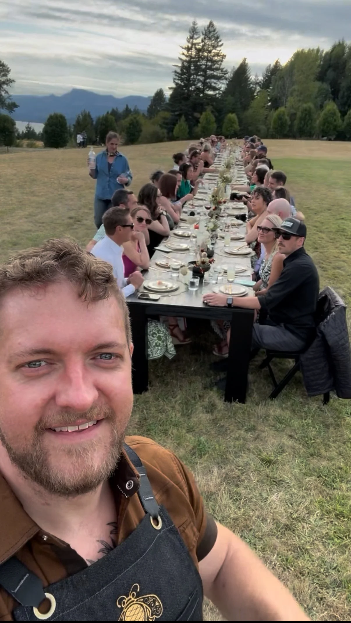 A man in the foreground taking a selfie at a long outdoor dinner event on a grassy field with mountains and trees in the background. Guests are seated along both sides of the long table, with plates, glasses, and floral centerpieces.