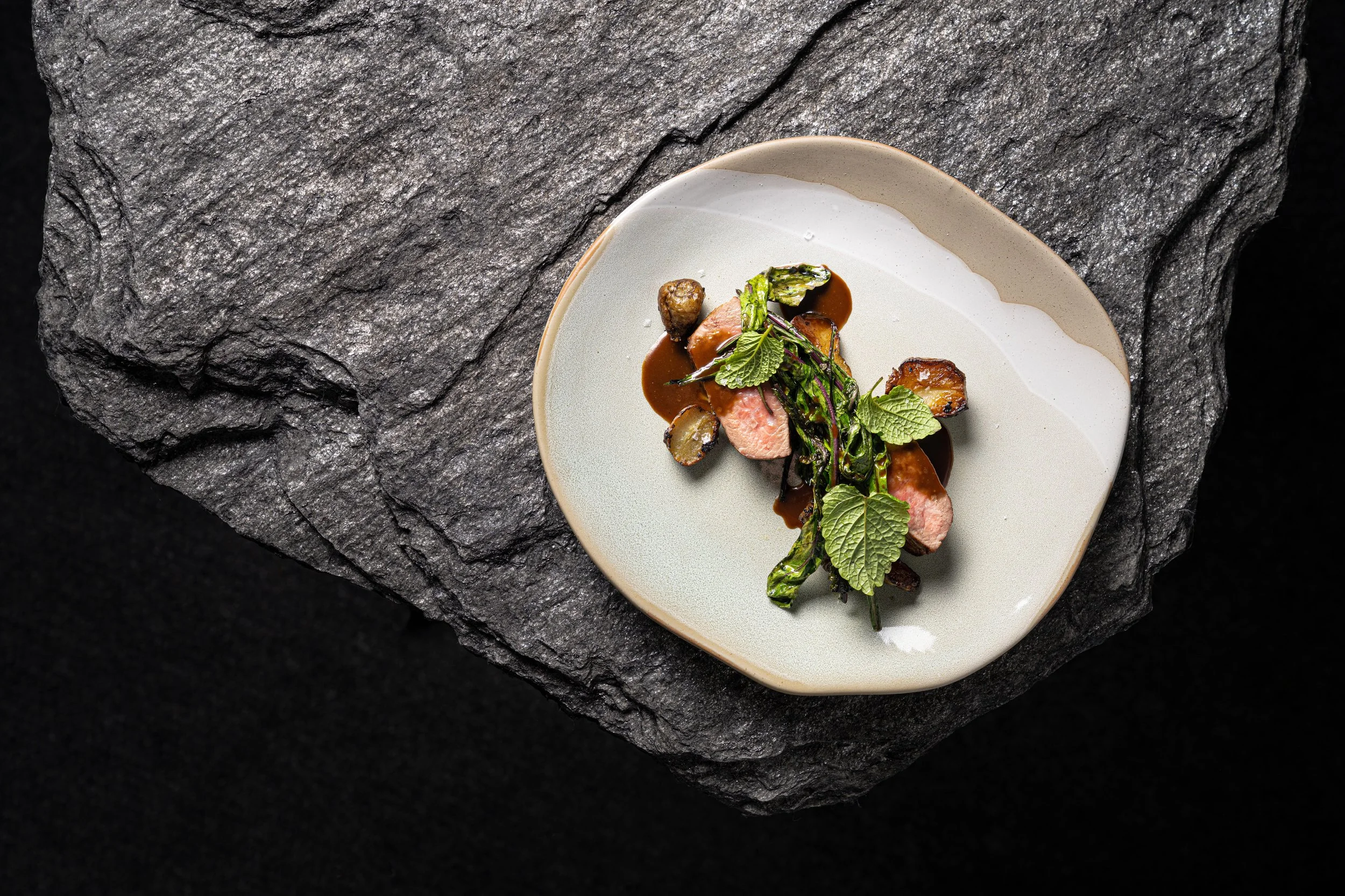 A gourmet dish with sliced pink meat, green leafy herbs, and roasted vegetables drizzled with sauce on a white plate against a textured dark stone background.