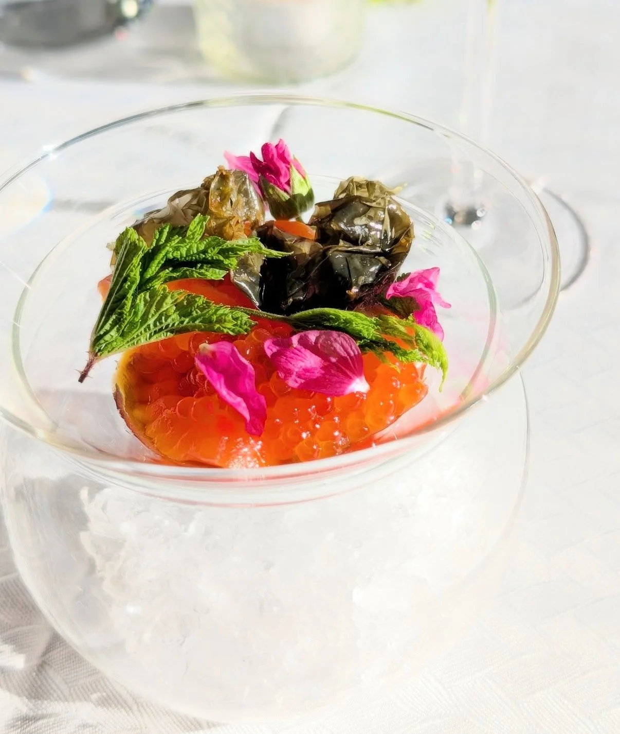 A clear glass bowl contains salmon roe, green herbs, pink flower petals, and black wrapped items, garnished with pink flower petals and green leaves.