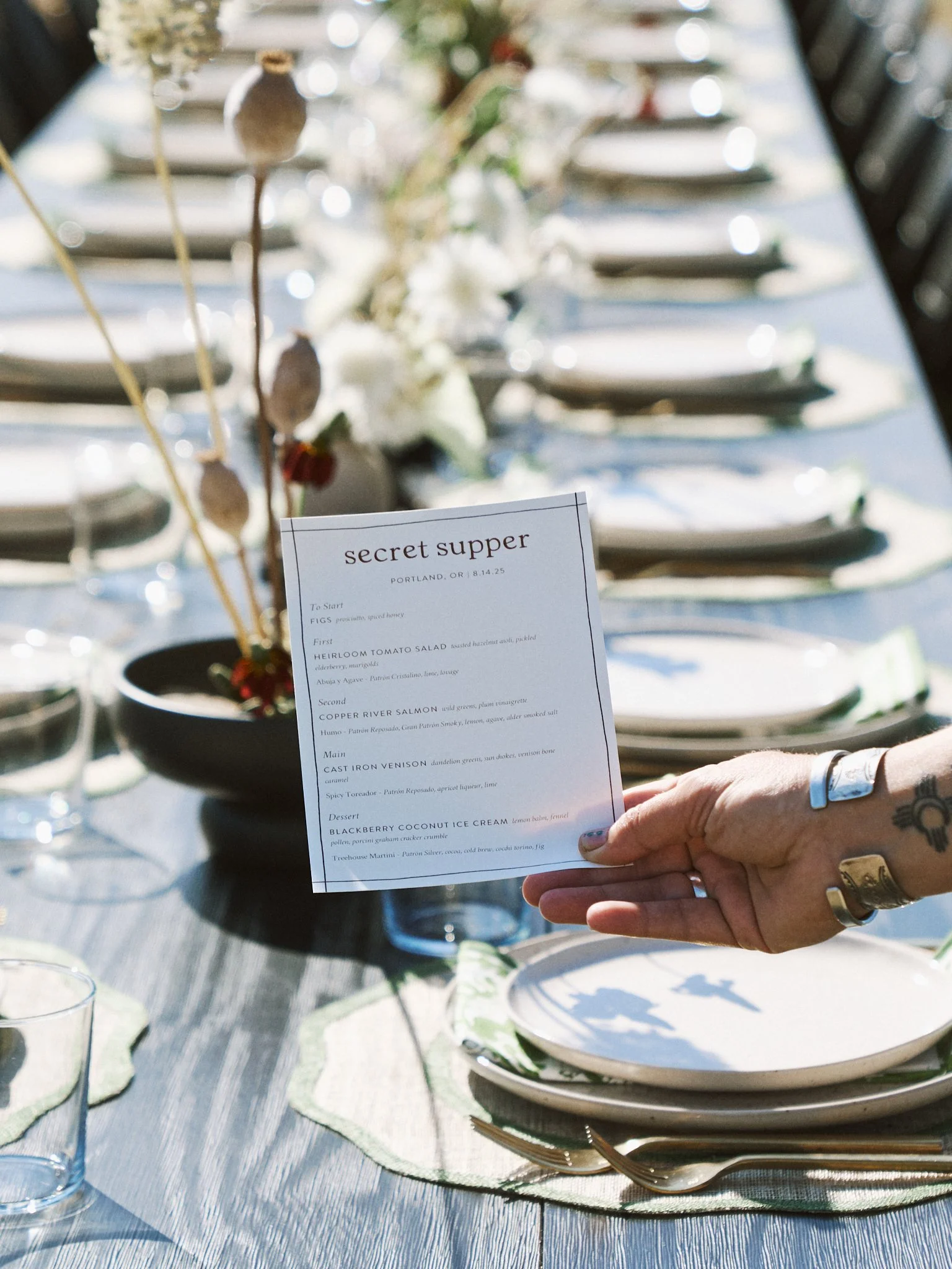 Person holding a menu at a formal outdoor dinner table set with plates, glasses, and napkins, decorated with flowers and greenery.