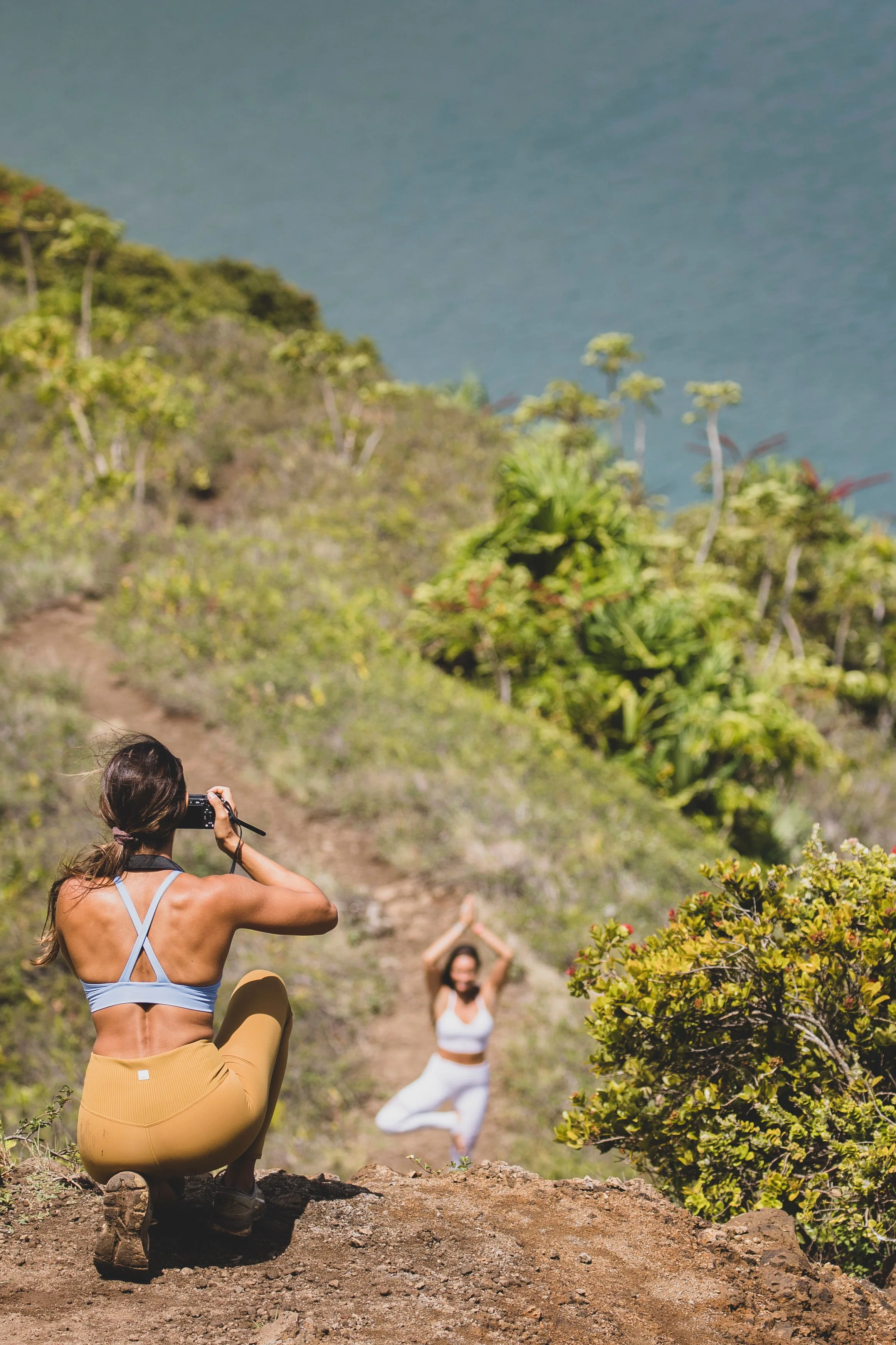 A woman in yellow leggings and a sports bra kneeling on a dirt trail taking a photo of another woman practicing yoga in a white outfit on a hillside surrounded by green plants, with water in the background.