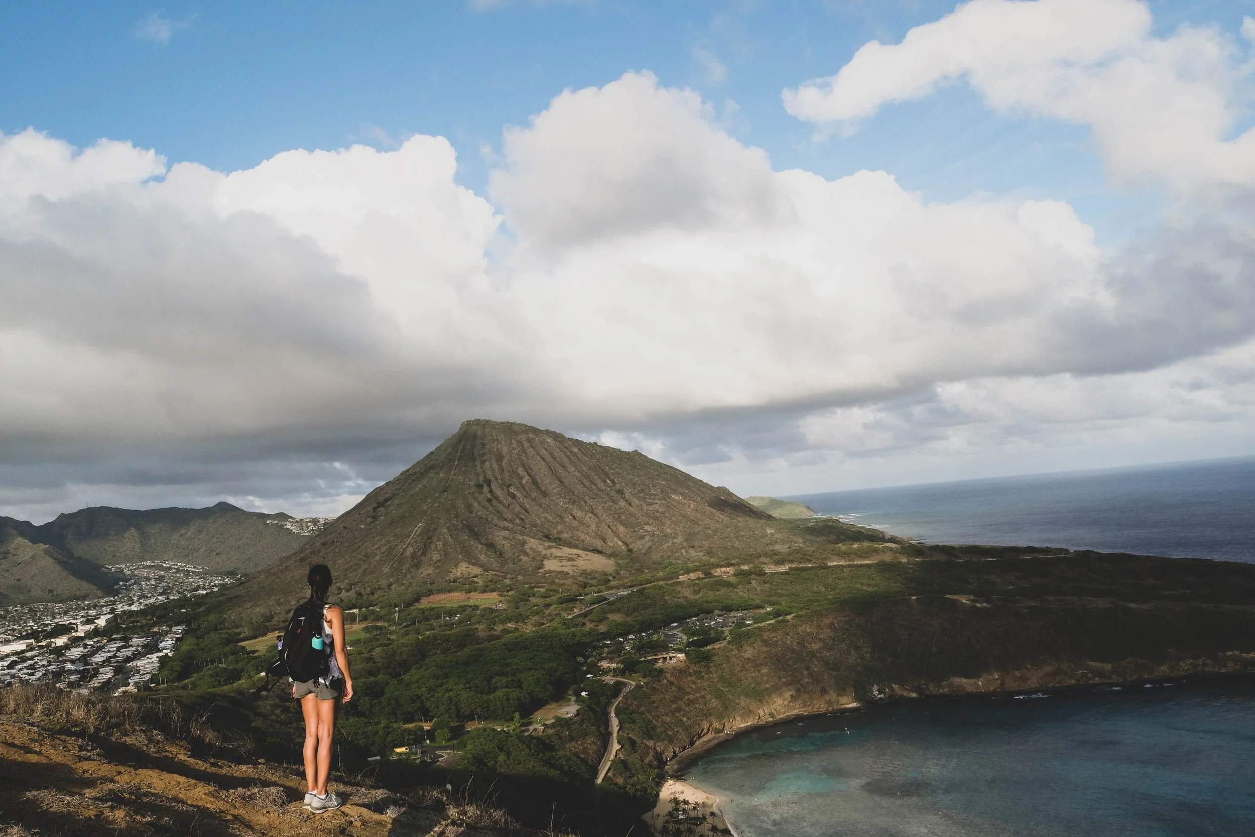 A woman with a backpack standing on a hill overlooking a mountain, the coast, and the ocean on a cloudy day.