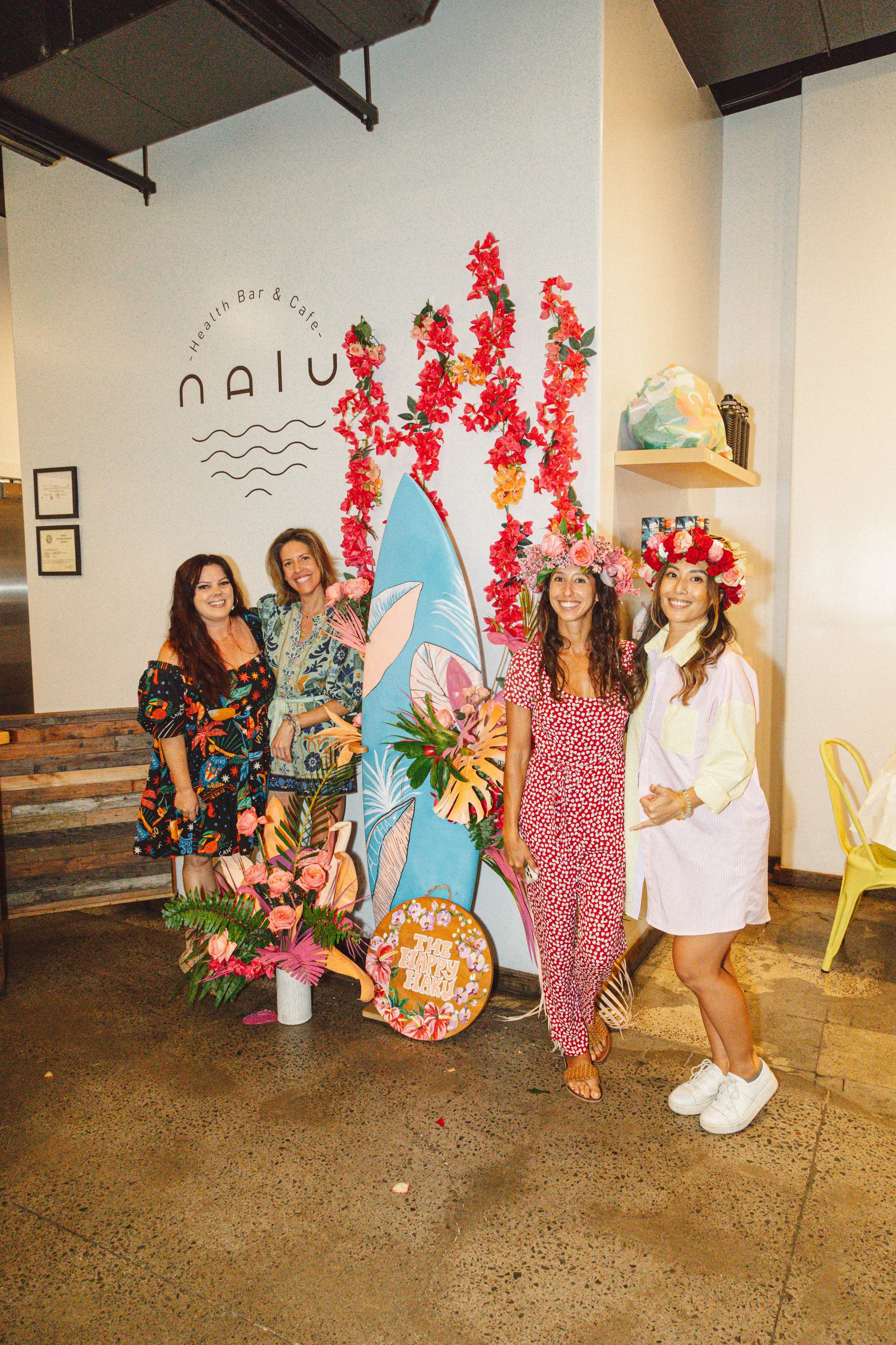 Four women standing inside a restaurant with floral decorations and a surfboard, wearing colorful clothing and flower crowns, smiling for the photo.