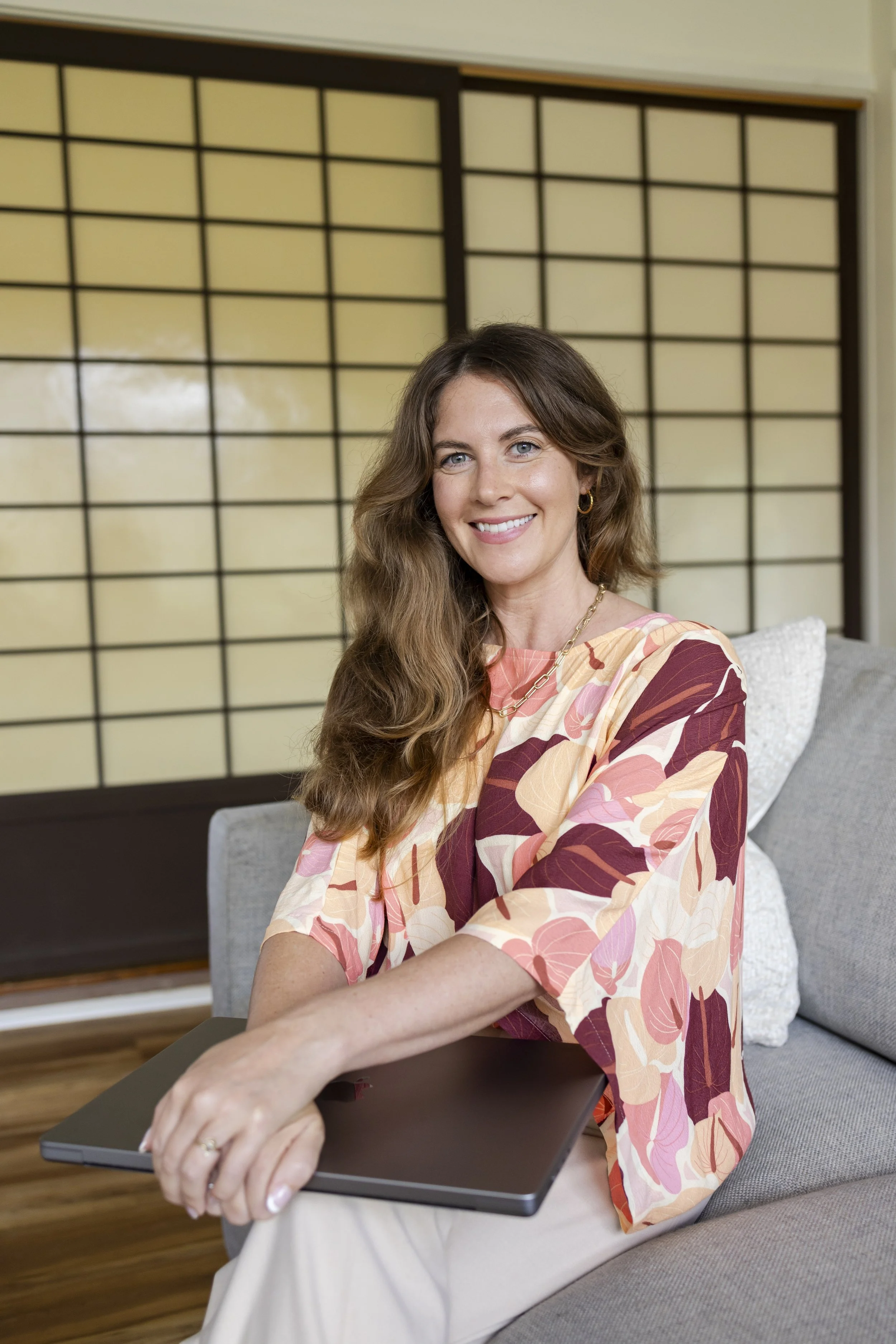 A woman with long wavy brown hair, smiling, sitting on a gray sofa in front of a shoji screen, wearing a multicolored floral blouse and holding a closed laptop on her lap.