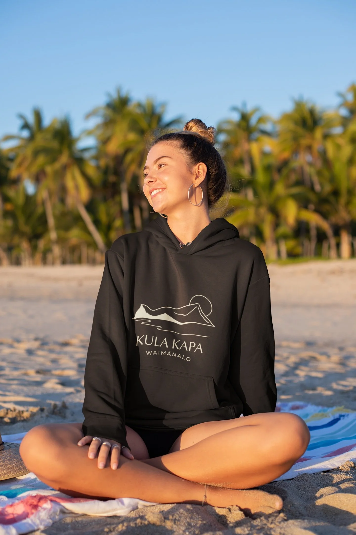 A young woman sitting cross-legged on a beach towel at sunset, wearing a black hoodie with white text and a mountain logo, smiling and looking to the side with palm trees in the background.