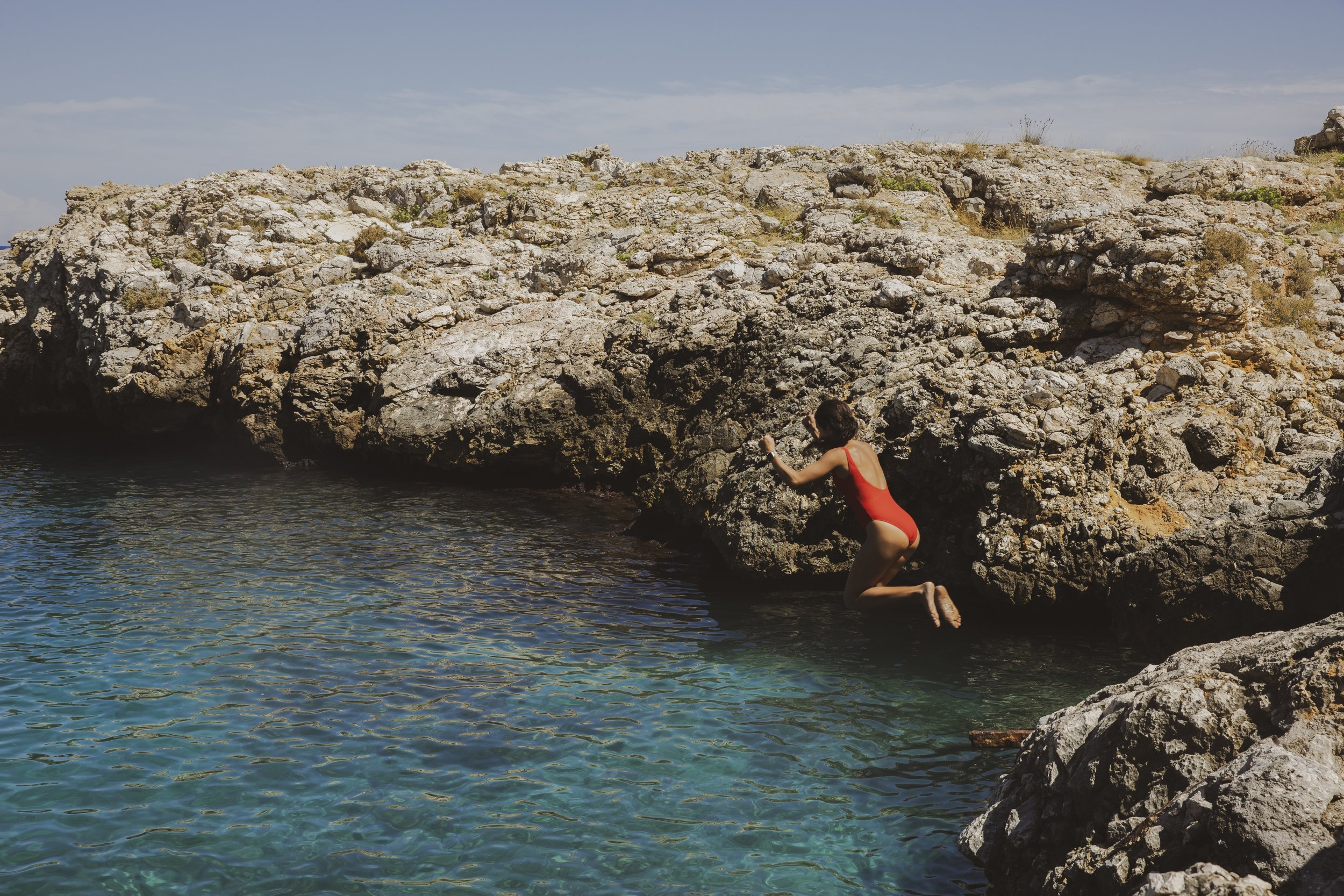A person in a red swimsuit jumping off a rocky ledge into a body of water near a rocky shoreline under a partly cloudy sky.