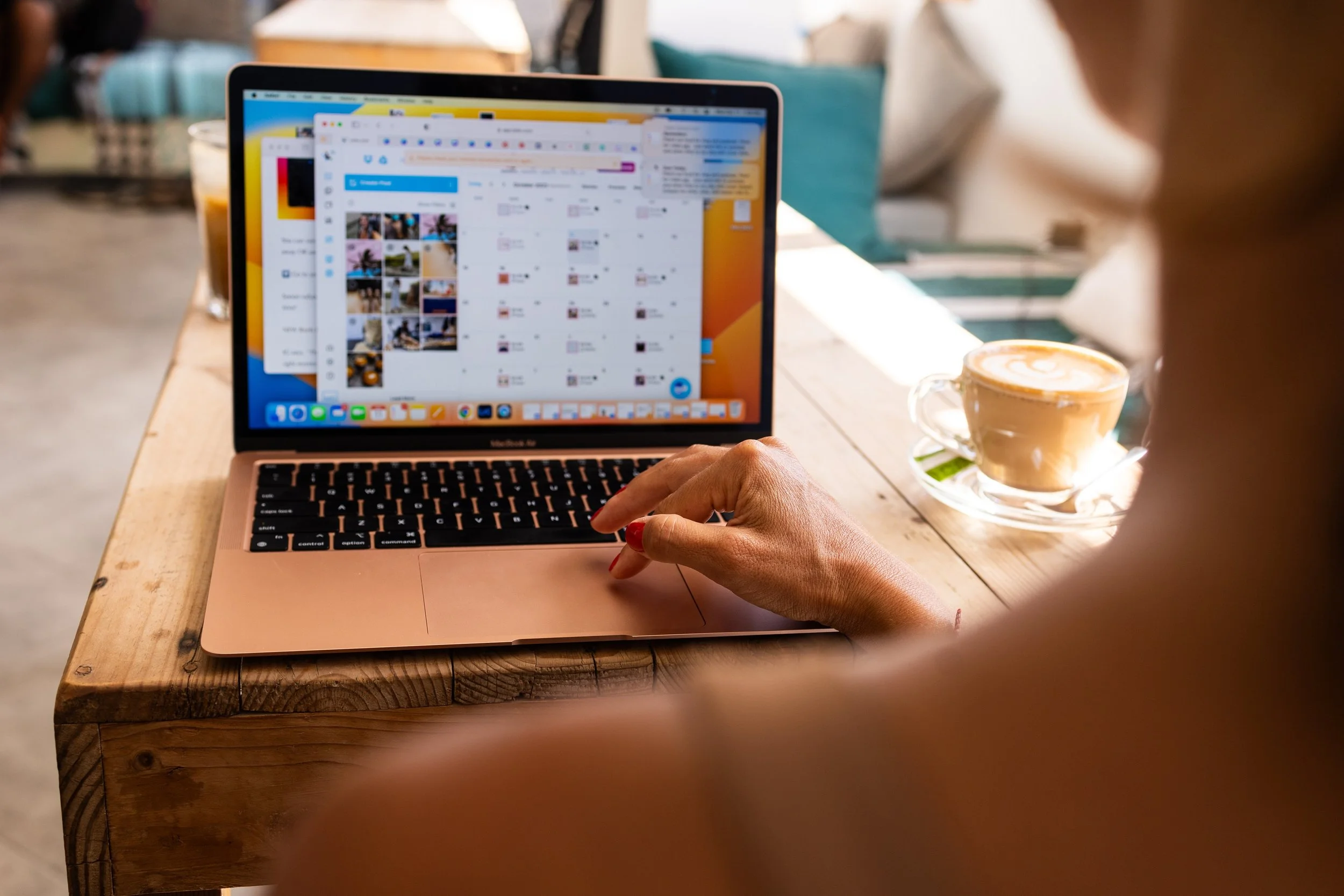 A person working on a pink MacBook with multiple open tabs and applications on a wooden table, next to a cup of coffee with latte art in a glass cup and saucer, in a cozy café setting.