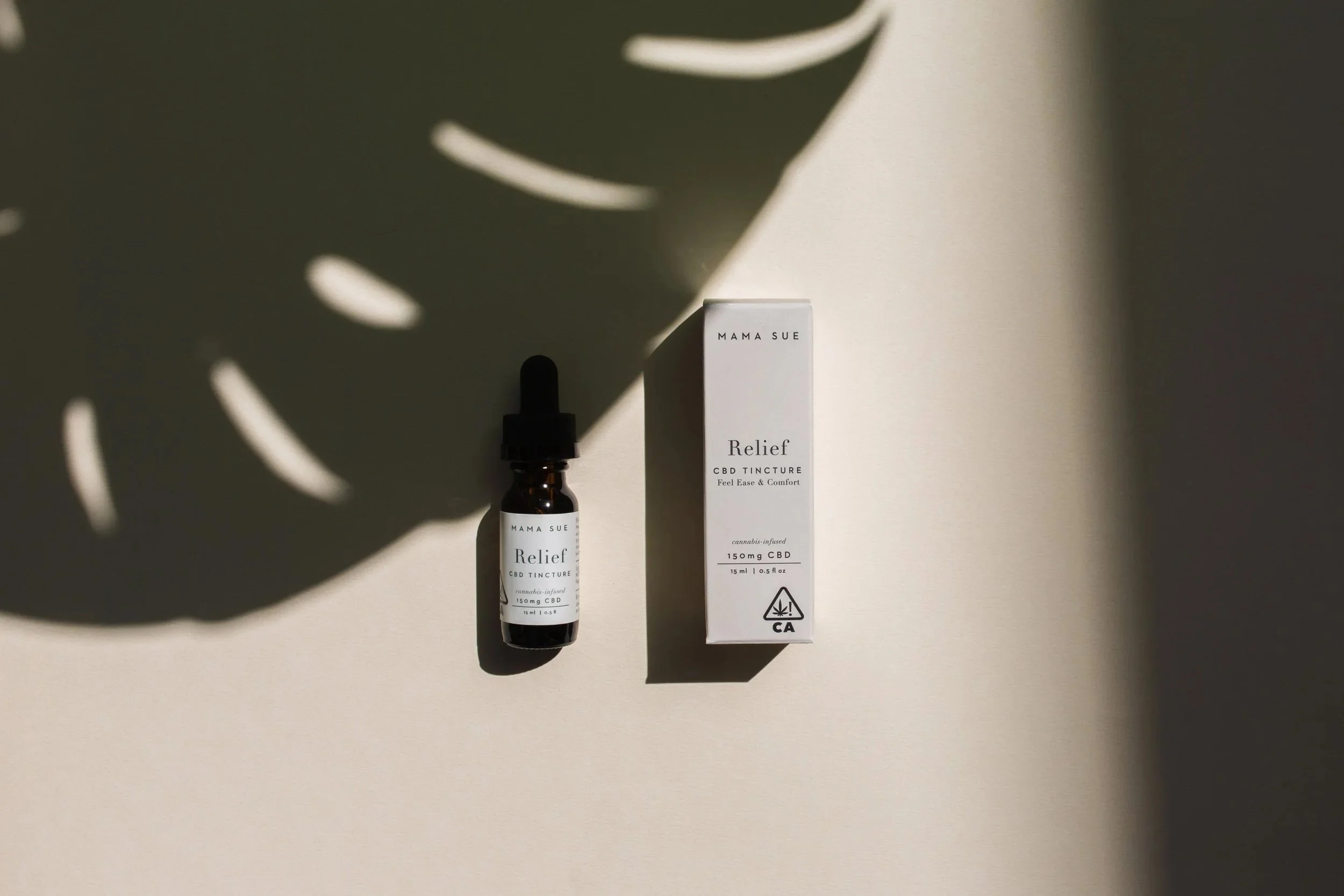 A small brown bottle with a black dropper cap and a white label, and a white rectangular box, both labeled 'Mama Sue Relief CBD Tincture,' positioned on a beige surface with shadows and light.