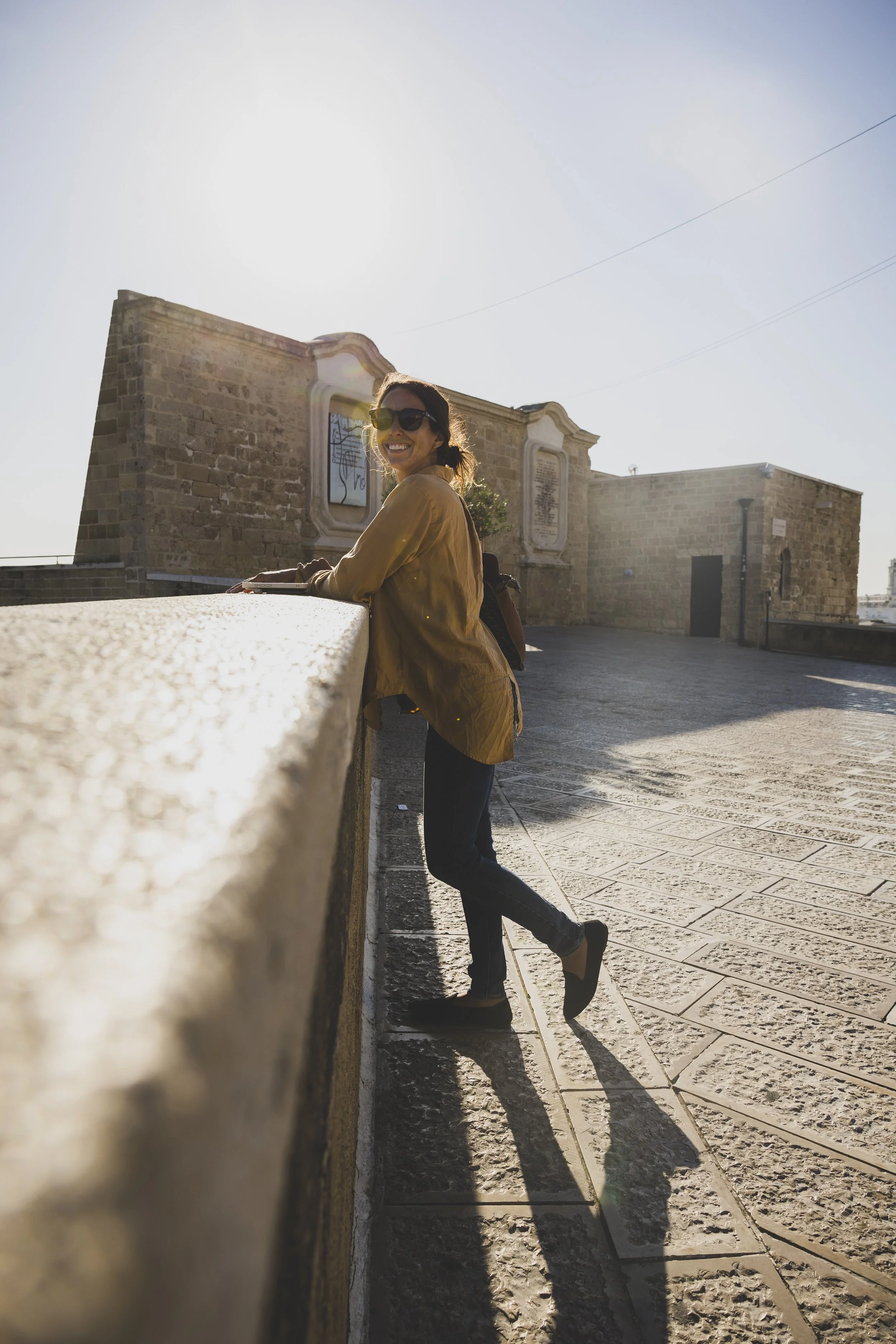 Woman smiling in sunglasses leaning on a stone wall on a sunny day.