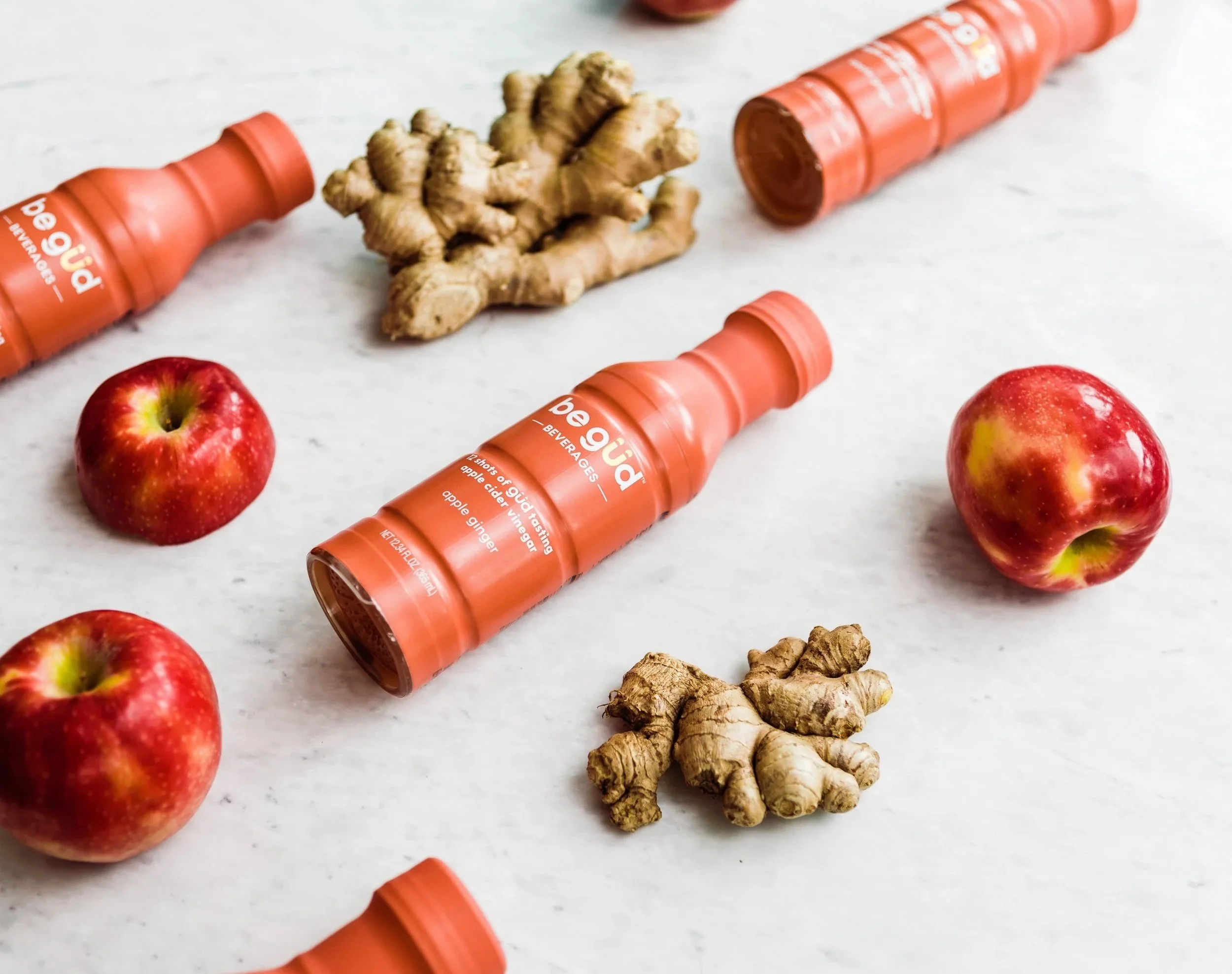 Three bottles of ginger apple vinegar drink labeled 'Pegü' on a white surface, along with three red apples and two pieces of ginger root.