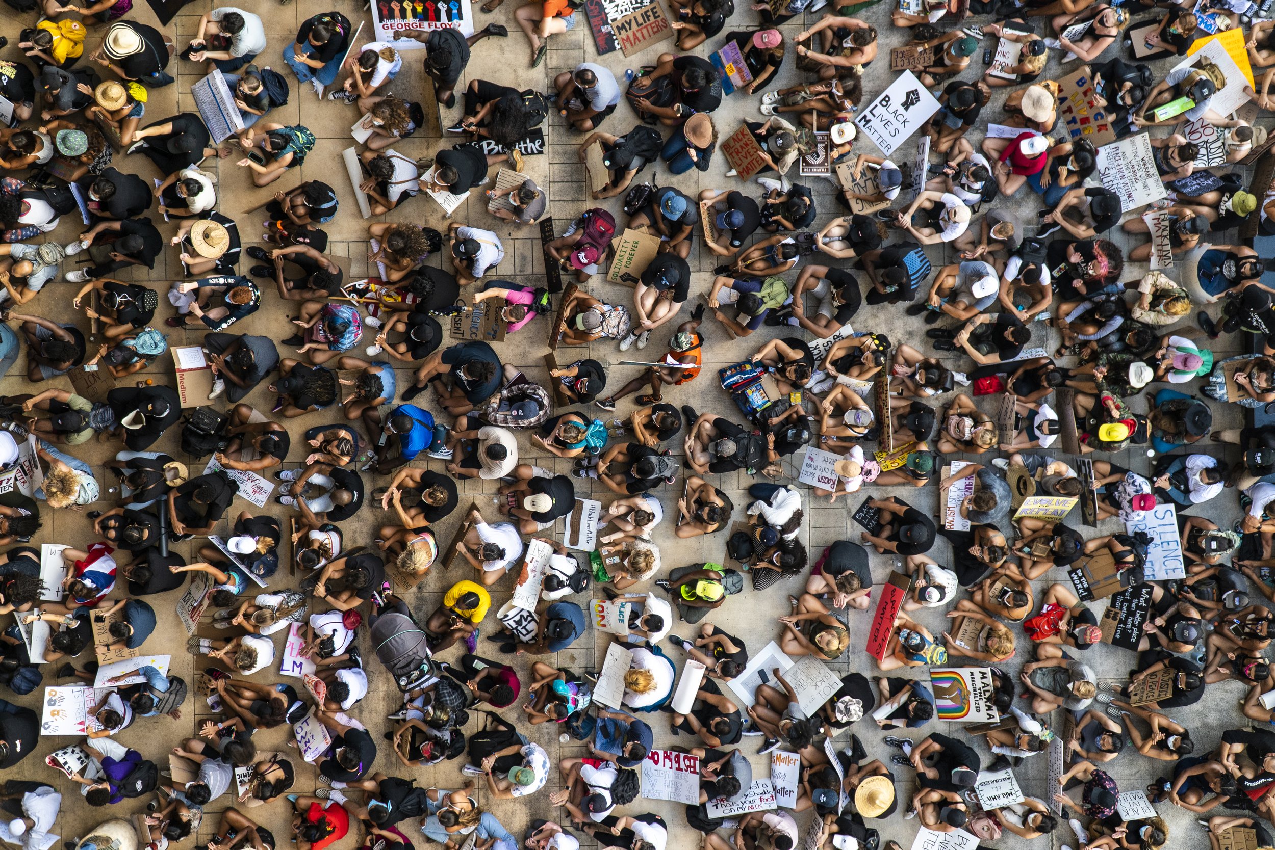 Aerial view of a large crowd of people sitting on the ground during a protest or rally, holding signs with messages supporting Black Lives Matter and social justice.