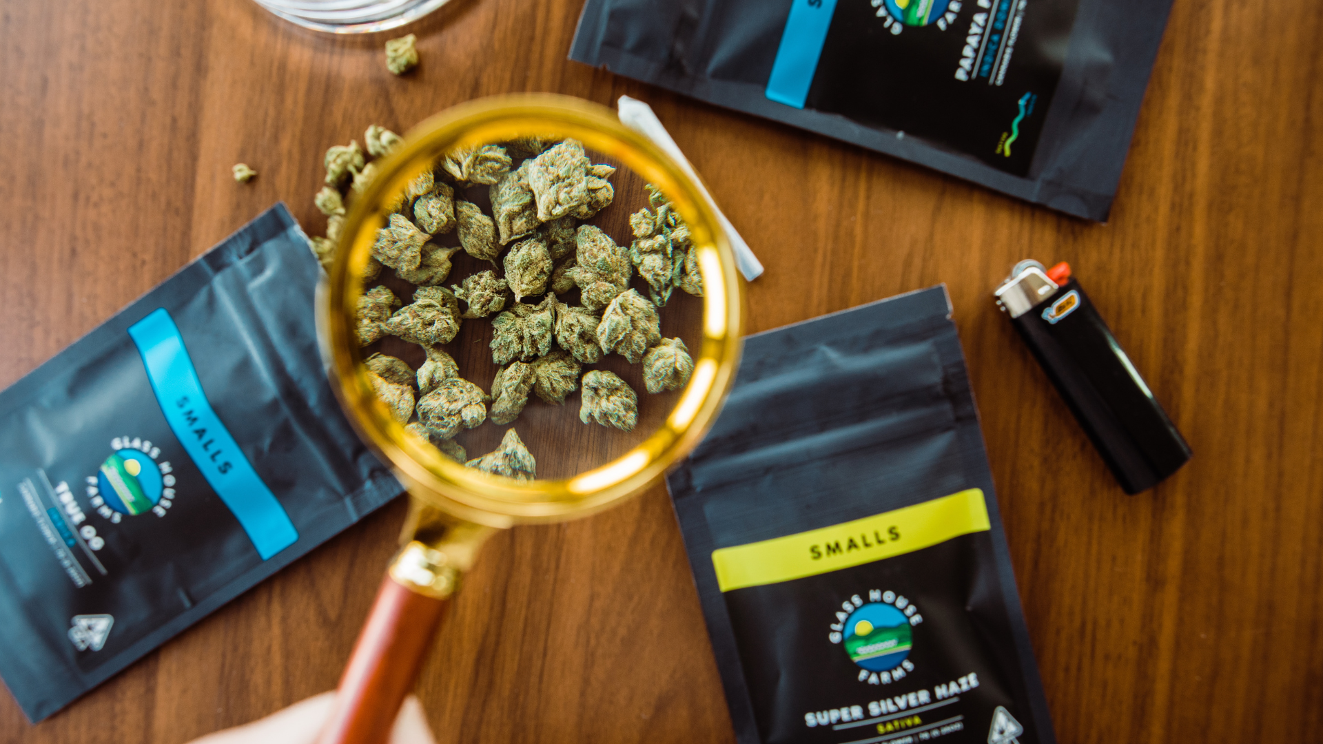 Various cannabis products in black packaging with a yellow label, a lighter, and a magnifying glass focusing on cannabis buds on a wooden surface.