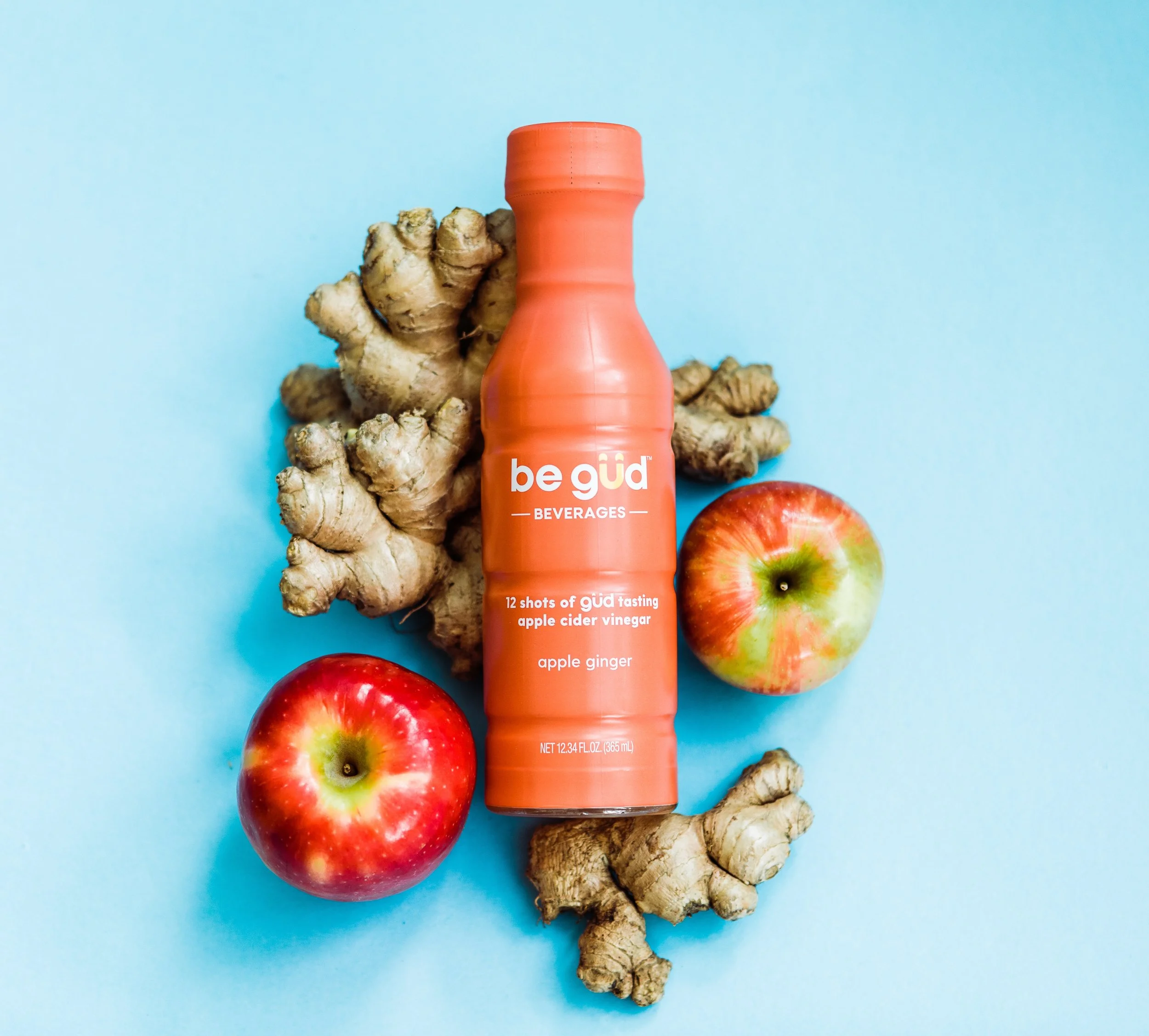 A bottle of be gúd apple cider vinegar tonic surrounded by ginger roots and two apples on a light blue background.