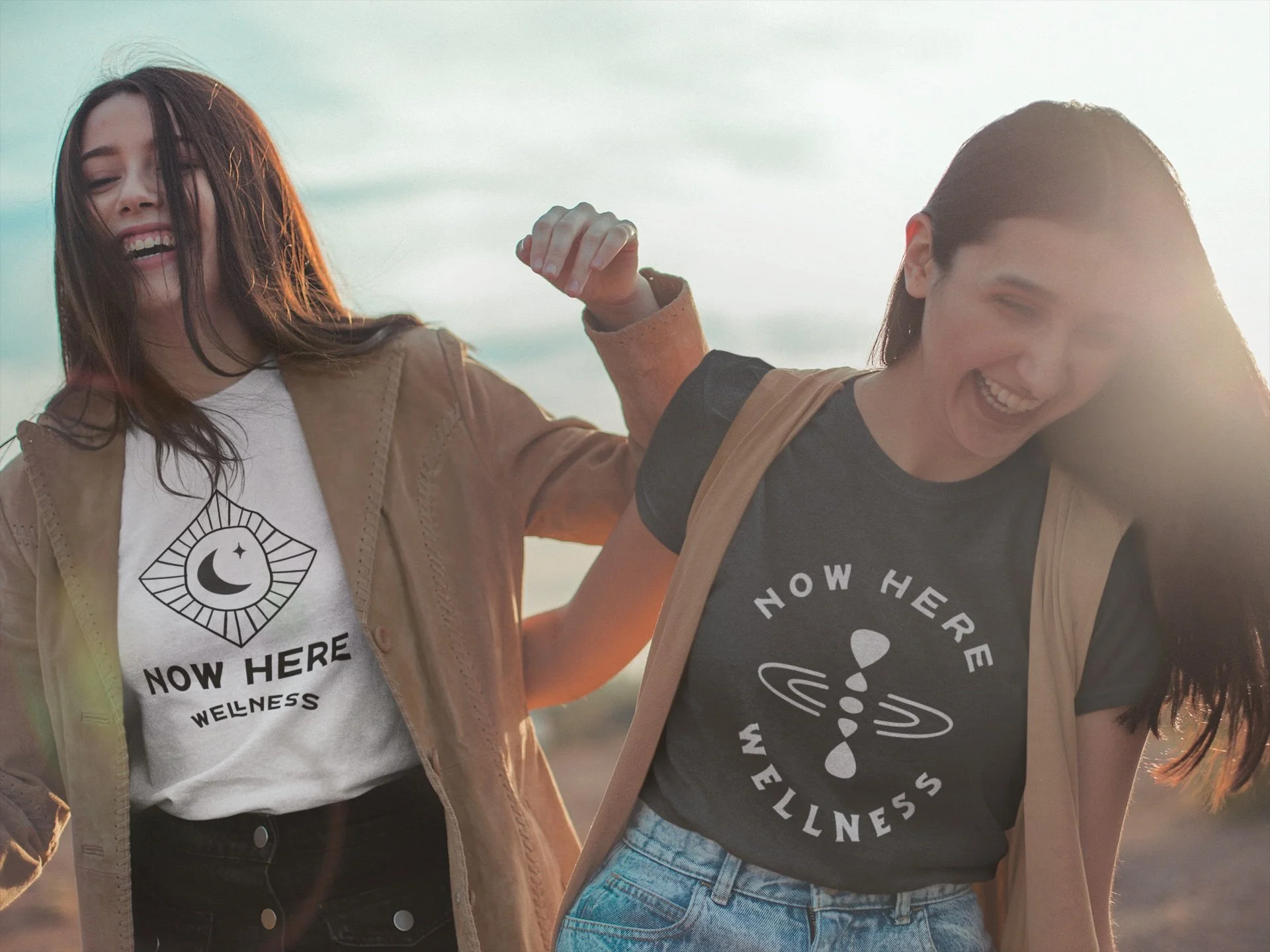 Two women smiling and enjoying themselves outdoors at sunset, wearing t-shirts with wellness-themed designs, one with a moon and stars and the other with an abstract pattern, arm-linking in a friendly pose.