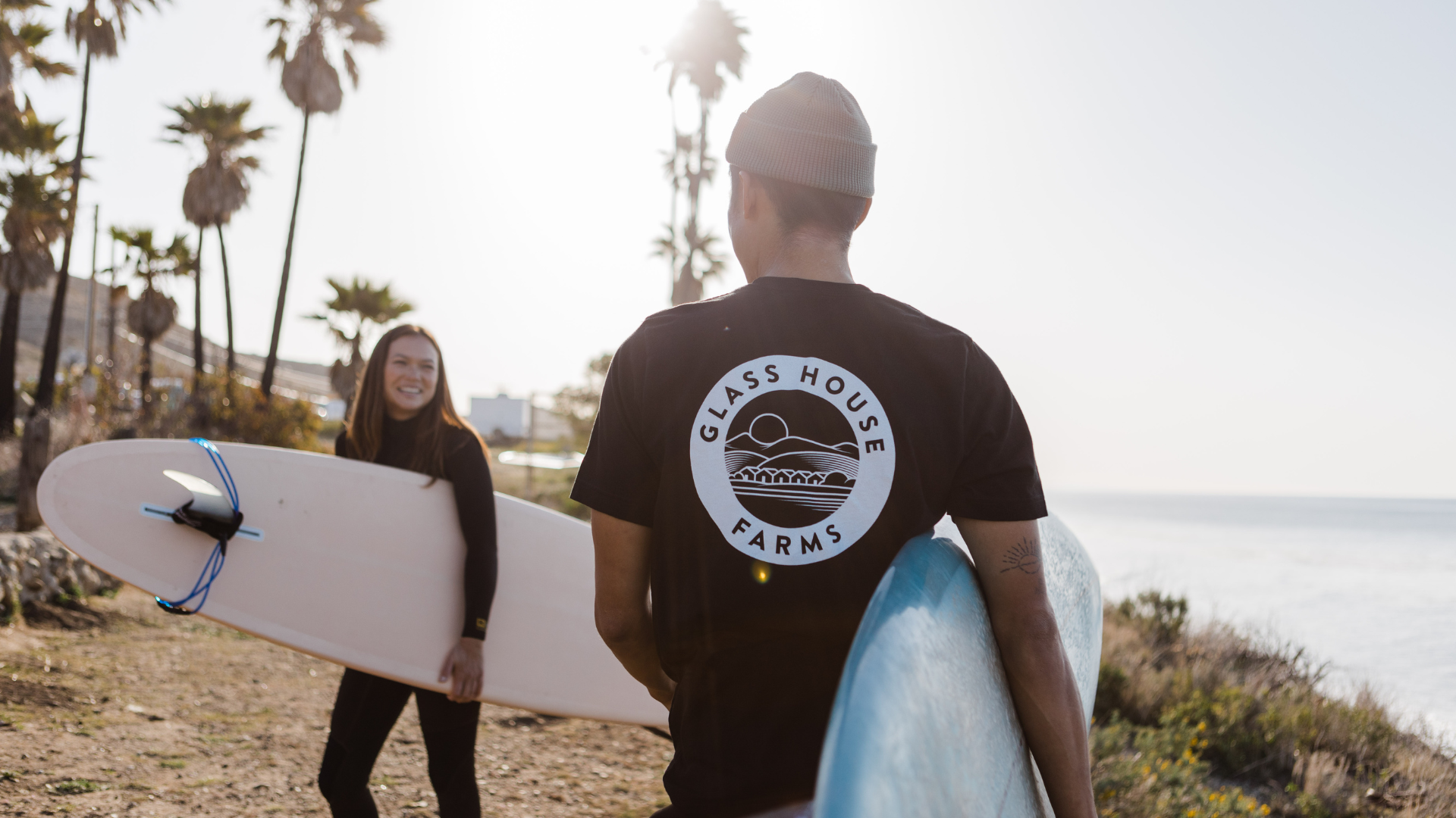 Two surfers on the beach, one man with a surfboard facing away, wearing a black T-shirt with 'Glass House Farms' logo, and a smiling woman holding her surfboard, palm trees, and ocean in the background.