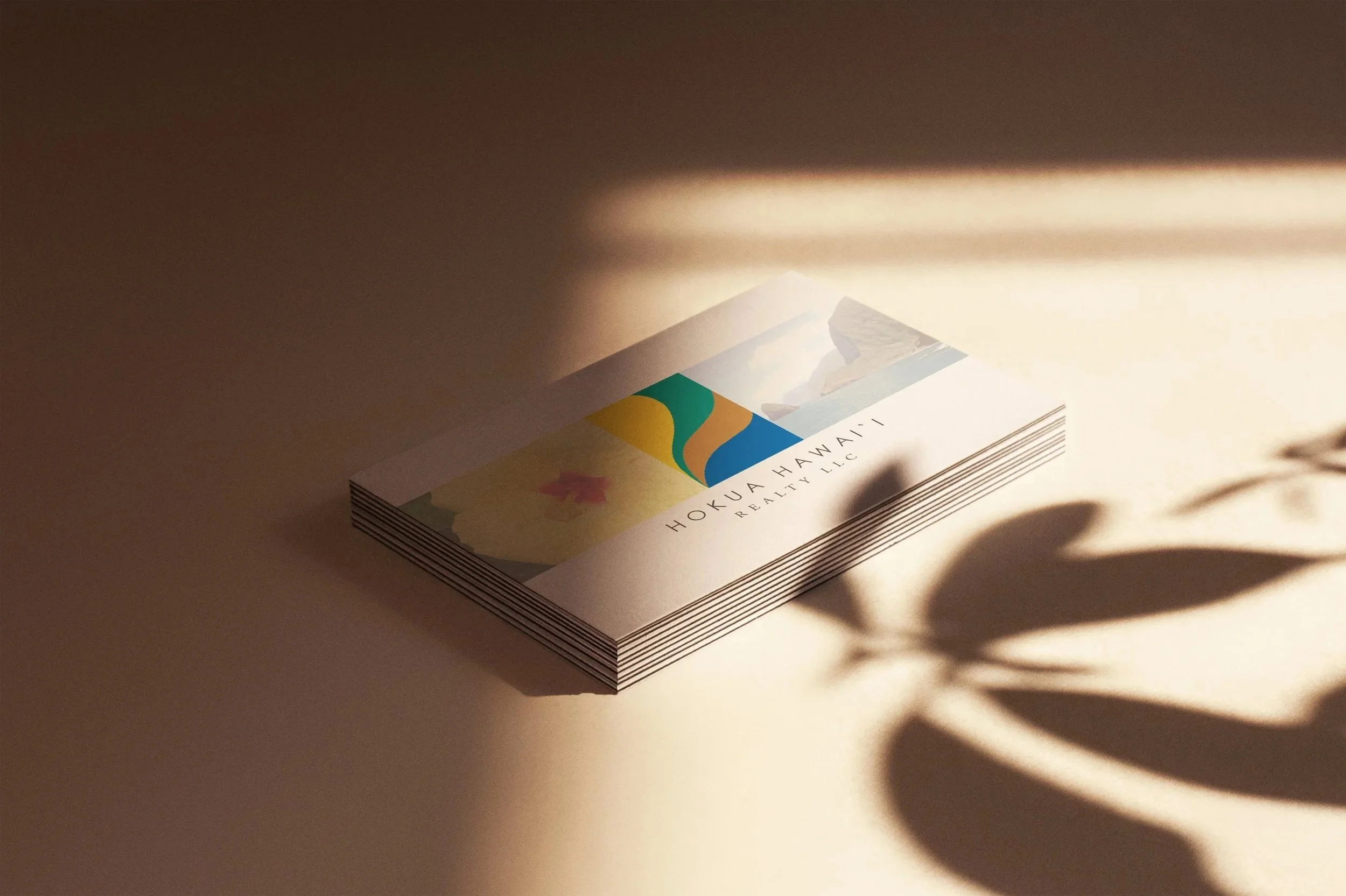 A stack of brochures or business cards with the text 'HOKUA HAWAII REALTY LLC' on the top one, featuring colorful abstract artwork and a mountain landscape image, with shadows and a leaf cast on a beige surface.