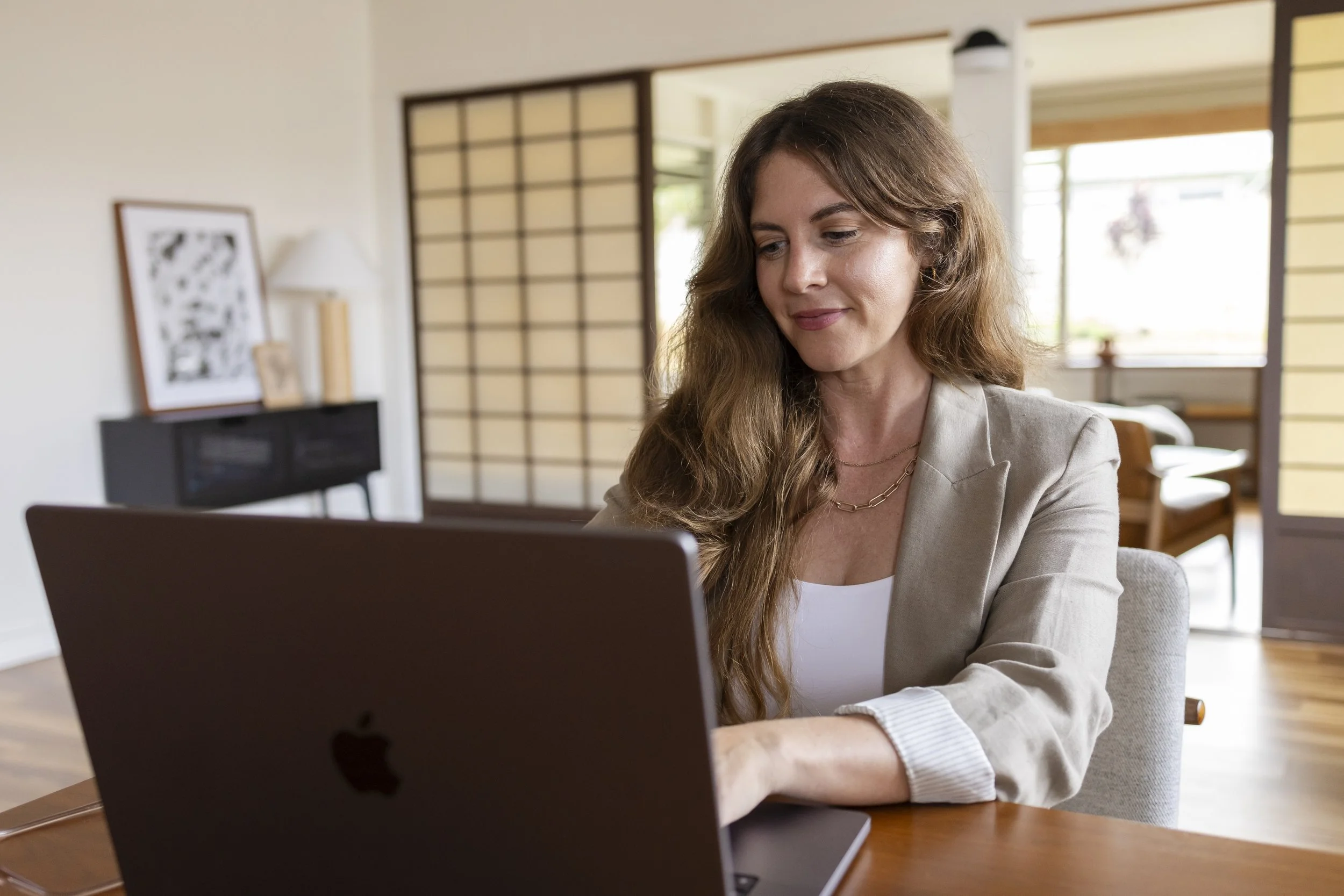 A woman with wavy brown hair and a light beige blazer sitting at a wooden desk in a bright, modern room, looking at a silver MacBook laptop.