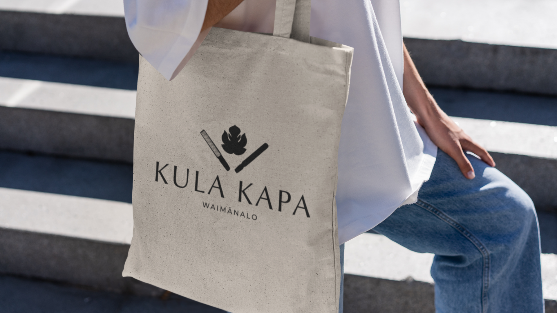 Person sitting on stairs holding a beige tote bag with the logo 'KULA KAPA WAIMANALO' and illustrations of a matchstick, a piece of fire, and a torch.