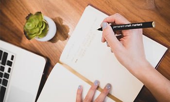 A person writing in a notebook with a black marker, with a laptop and a small potted plant nearby on a wooden desk.