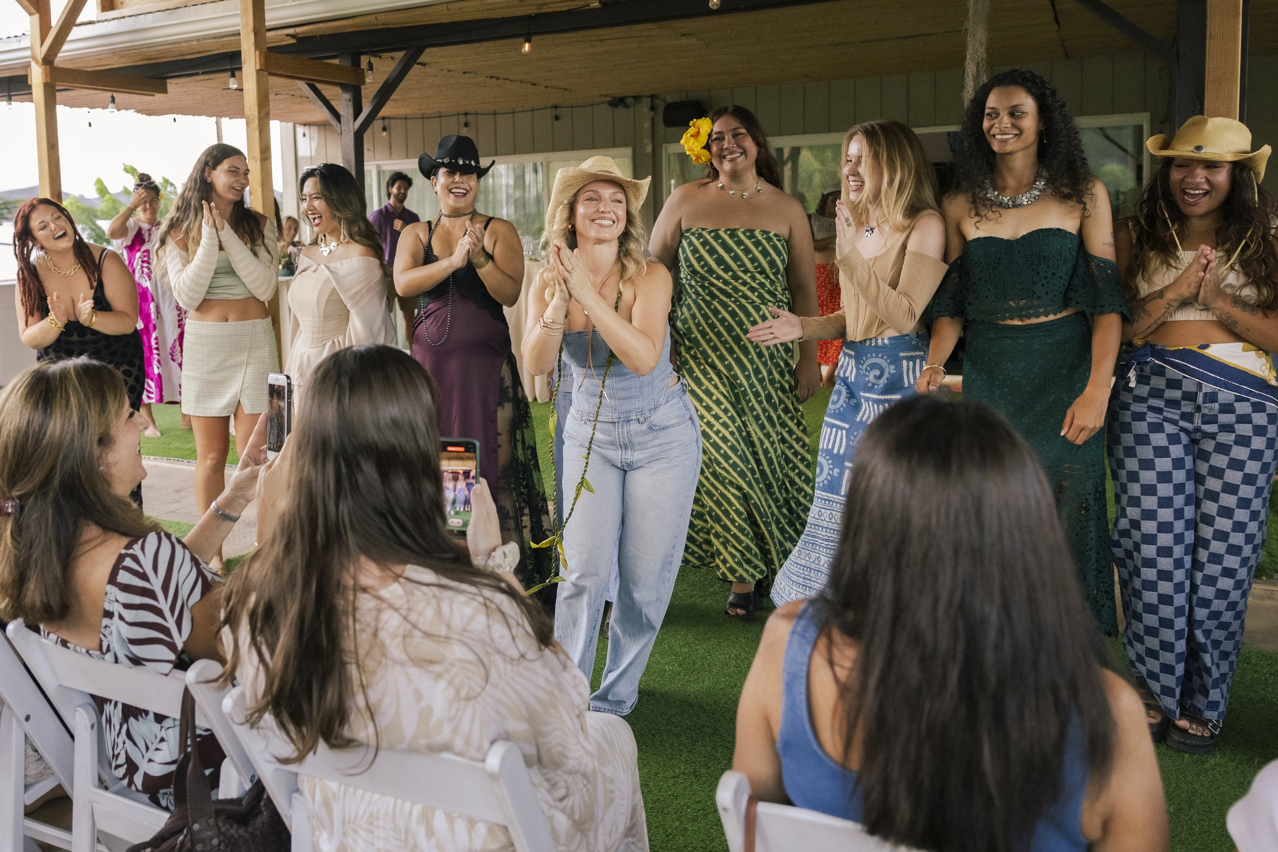 Group of women at a celebration, some in casual and bohemian style clothing, standing and smiling facing an audience with some taking pictures.