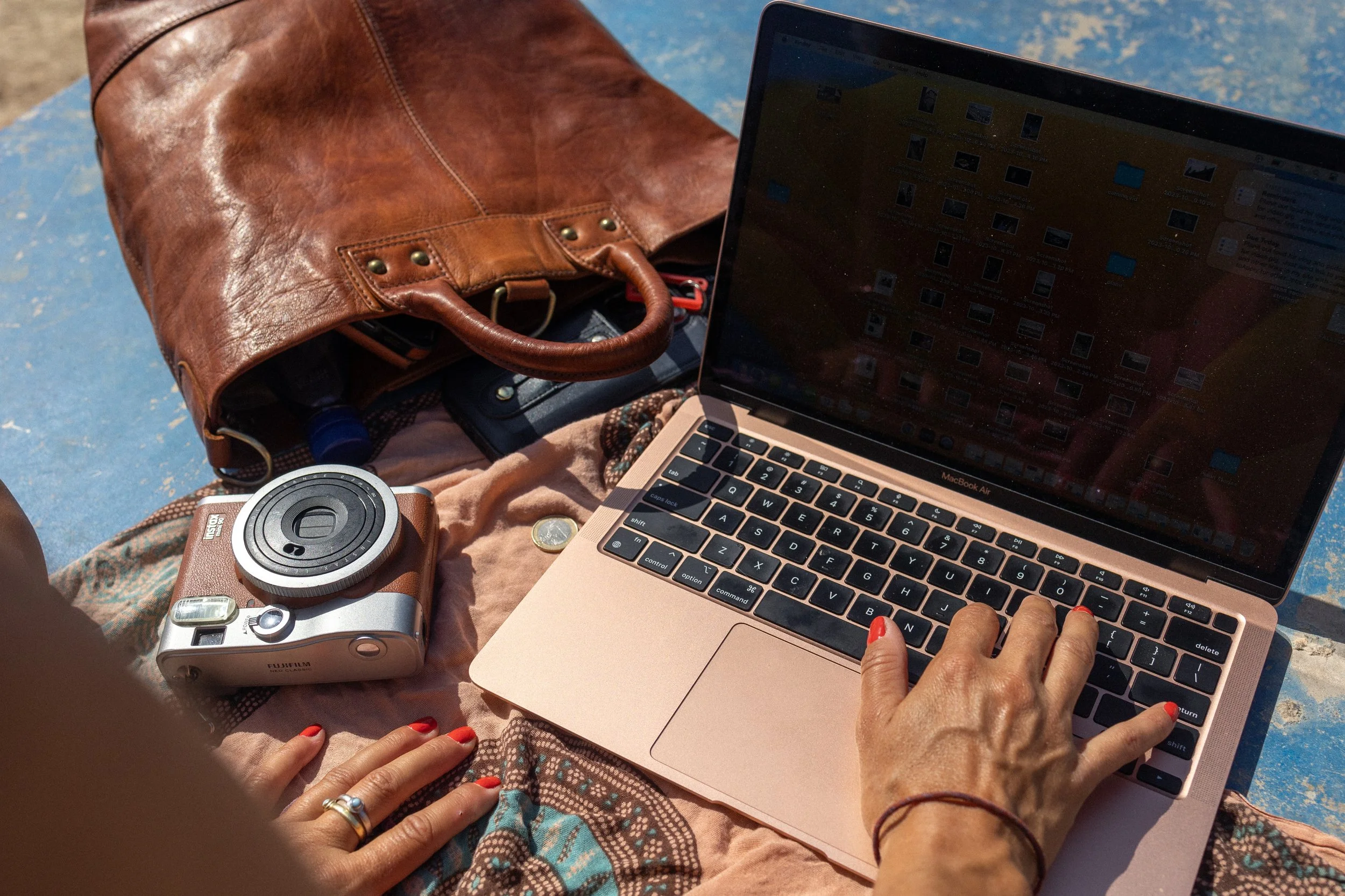 A person is typing on a MacBook Air laptop while sitting outdoors. There is a vintage camera, a leather bag, a coin, and a piece of cloth on the ground nearby.