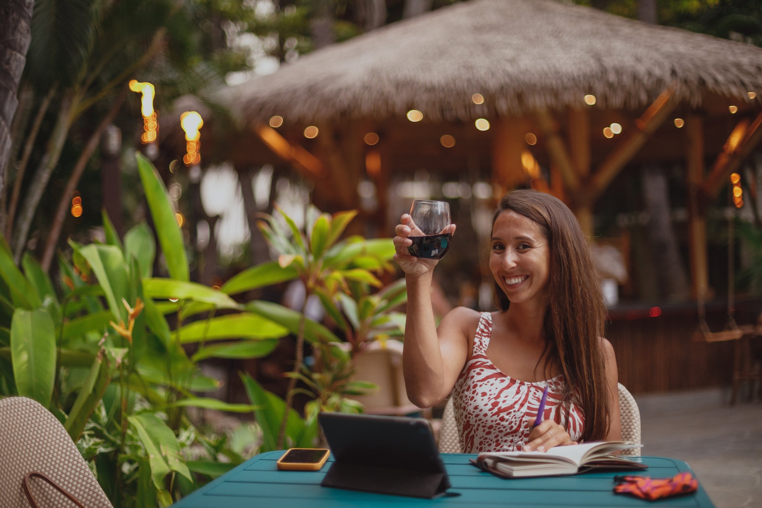 A woman sitting at a table outdoors at a tropical resort, smiling and holding a glass of red wine, with a smartphone, notebook, and pen on the table.
