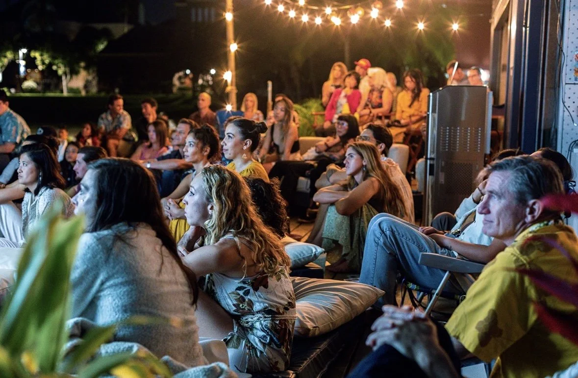 People sitting outdoors on cushions and chairs, attentively watching a performance or presentation at night, illuminated by string lights.