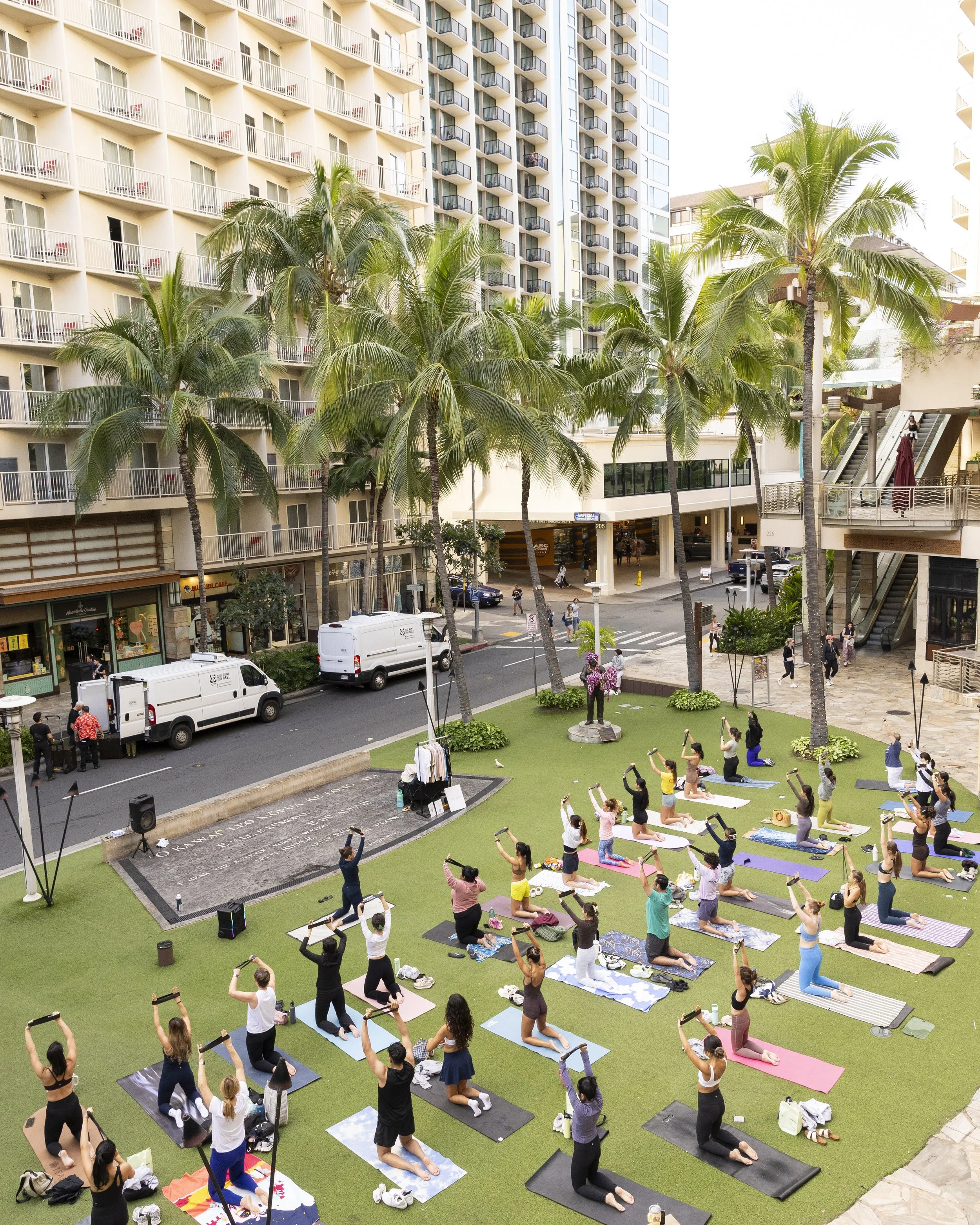 Group of people participating in an outdoor yoga class with some raising their arms, surrounded by palm trees and high-rise buildings in an urban setting.