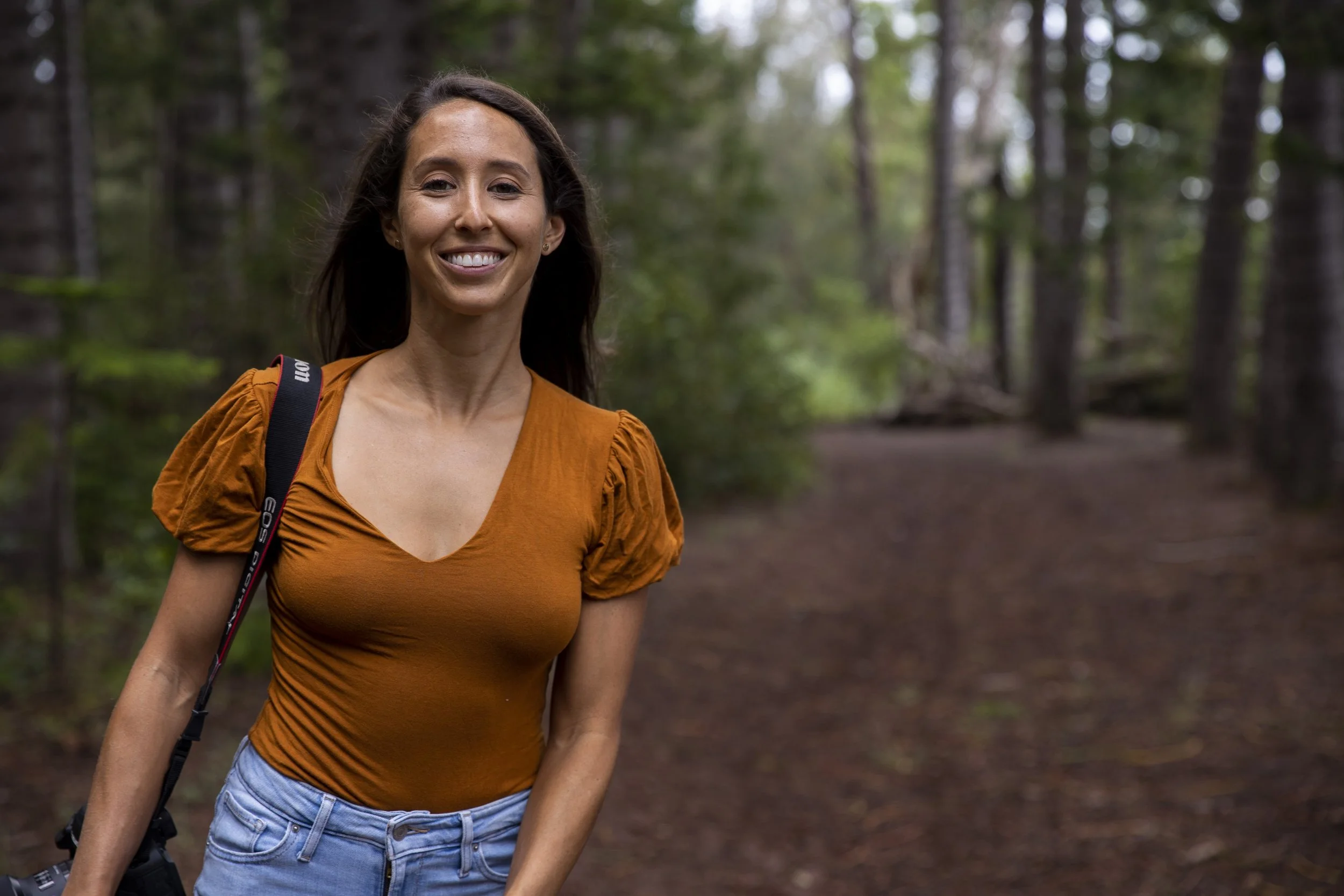 A smiling woman in a burnt orange top and light blue jeans standing in a wooded forest area with a camera strap over her shoulder and a camera in her hand.