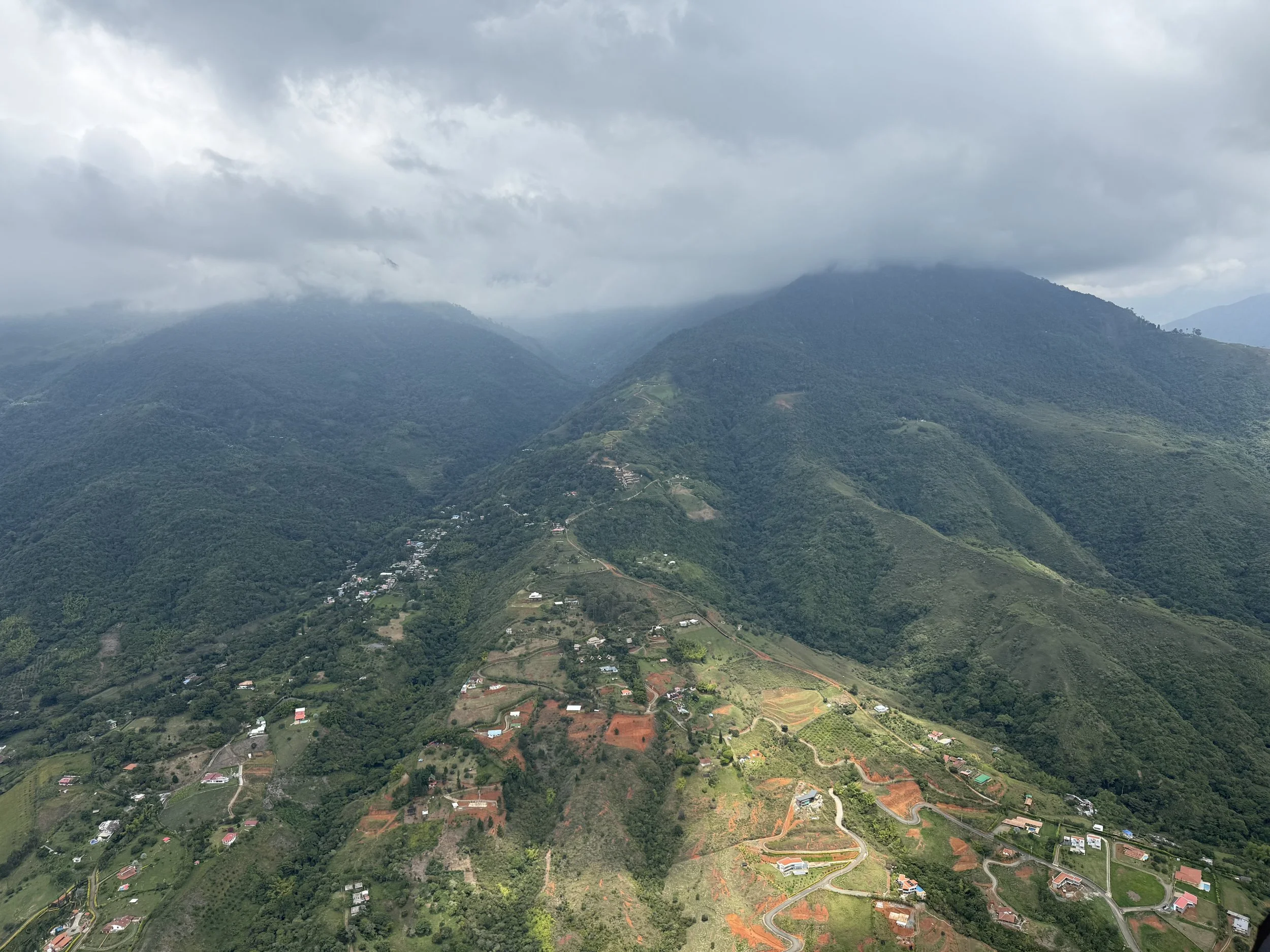 Aerial view of green mountain landscape with scattered houses, winding roads, and cloud-covered mountain peaks in the distance.