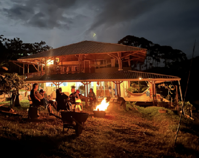 People sitting around a fire pit outside a two-story house at night, with dark clouds and trees in the background.