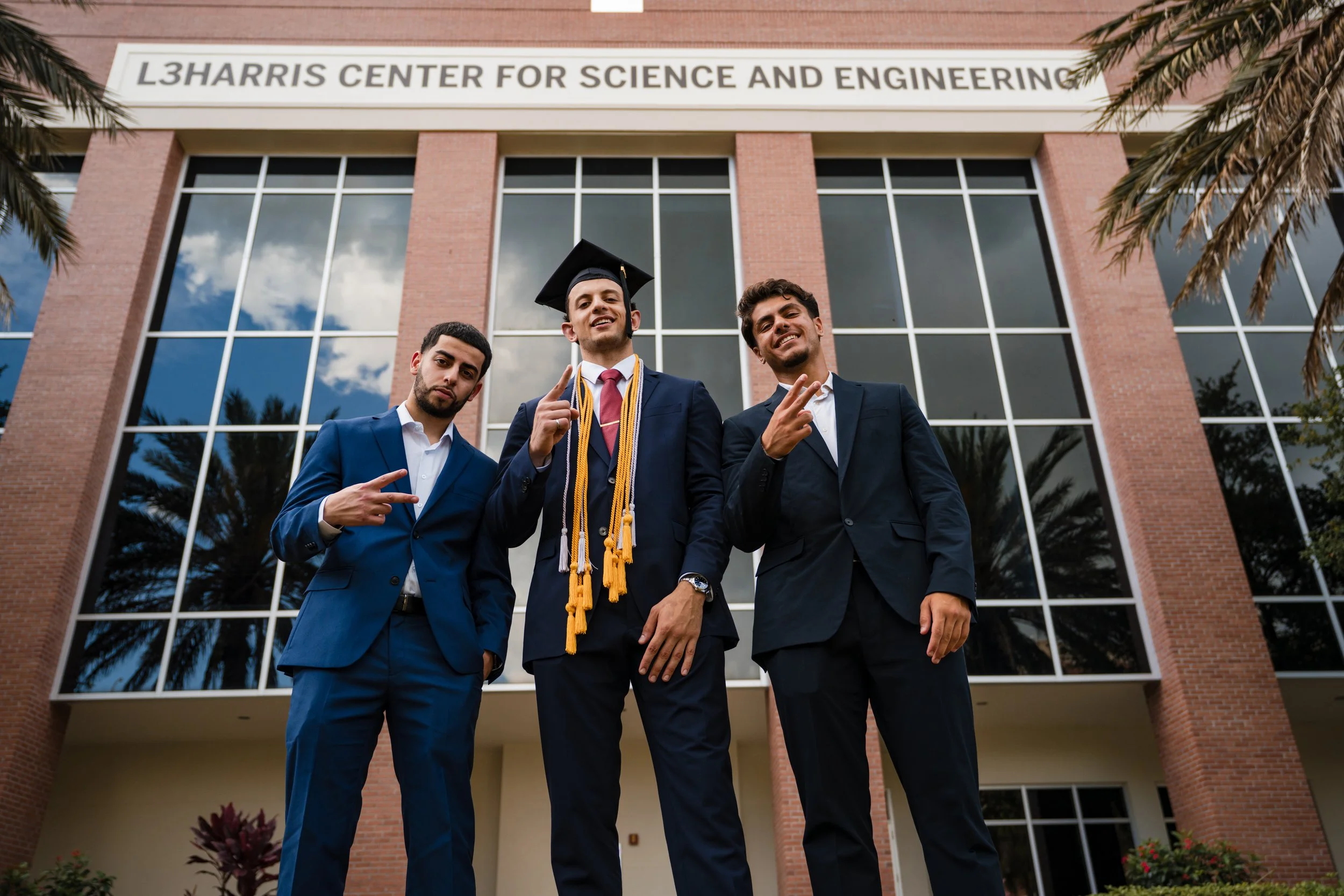 Florida Tech graduate poses with two friends in suits outside the L3Harris Center for Science and Engineering, proudly flashing hand signs in celebration.