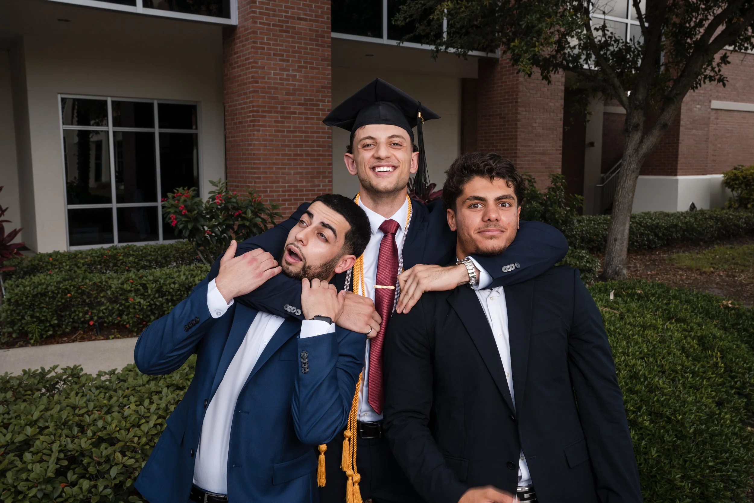 Florida Tech graduate in cap and gown laughs while playfully putting two friends in headlocks during a fun graduation photo outside the campus building.