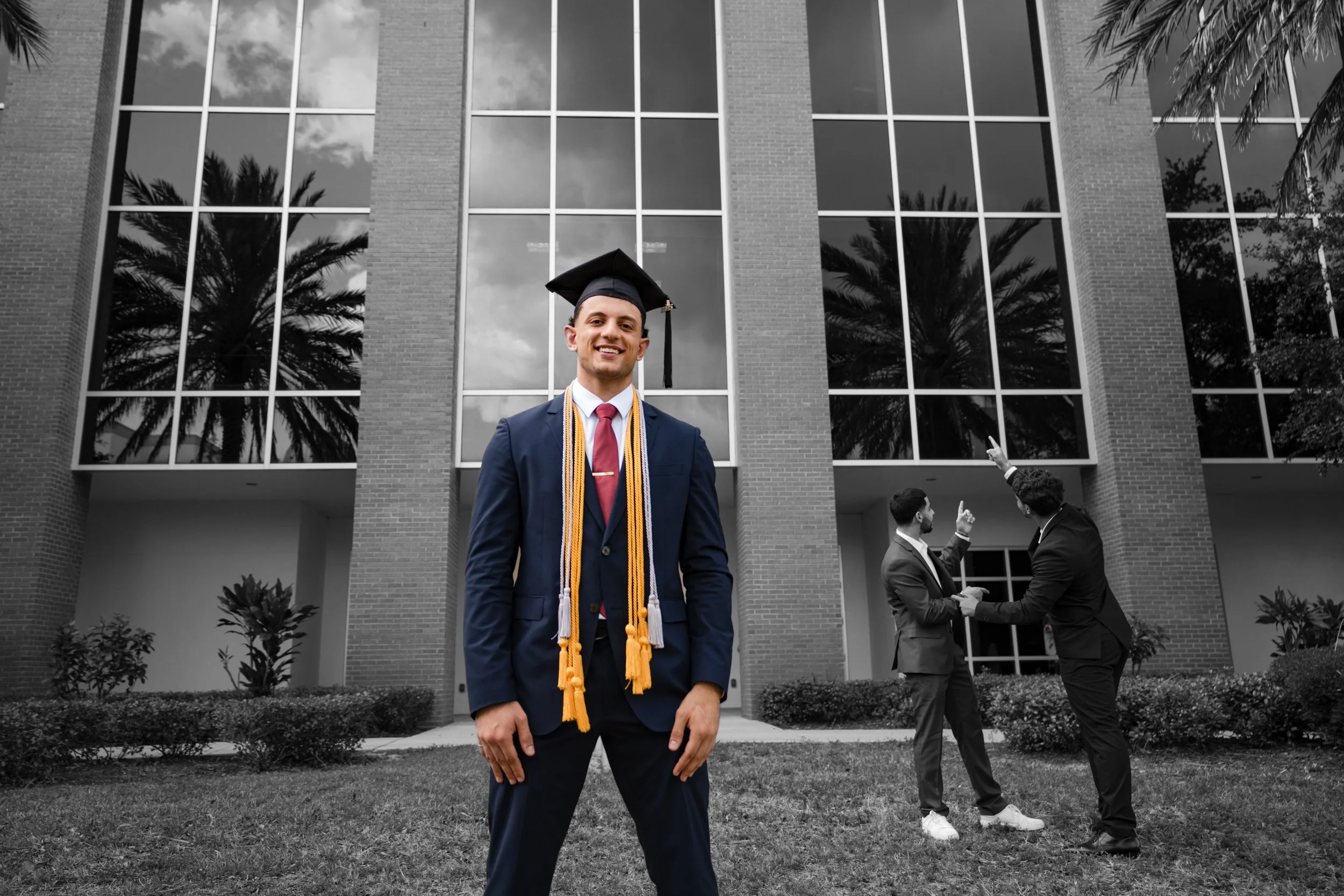 College graduate in color stands confidently in front of a modern glass building while friends in black and white gesture playfully in the background.