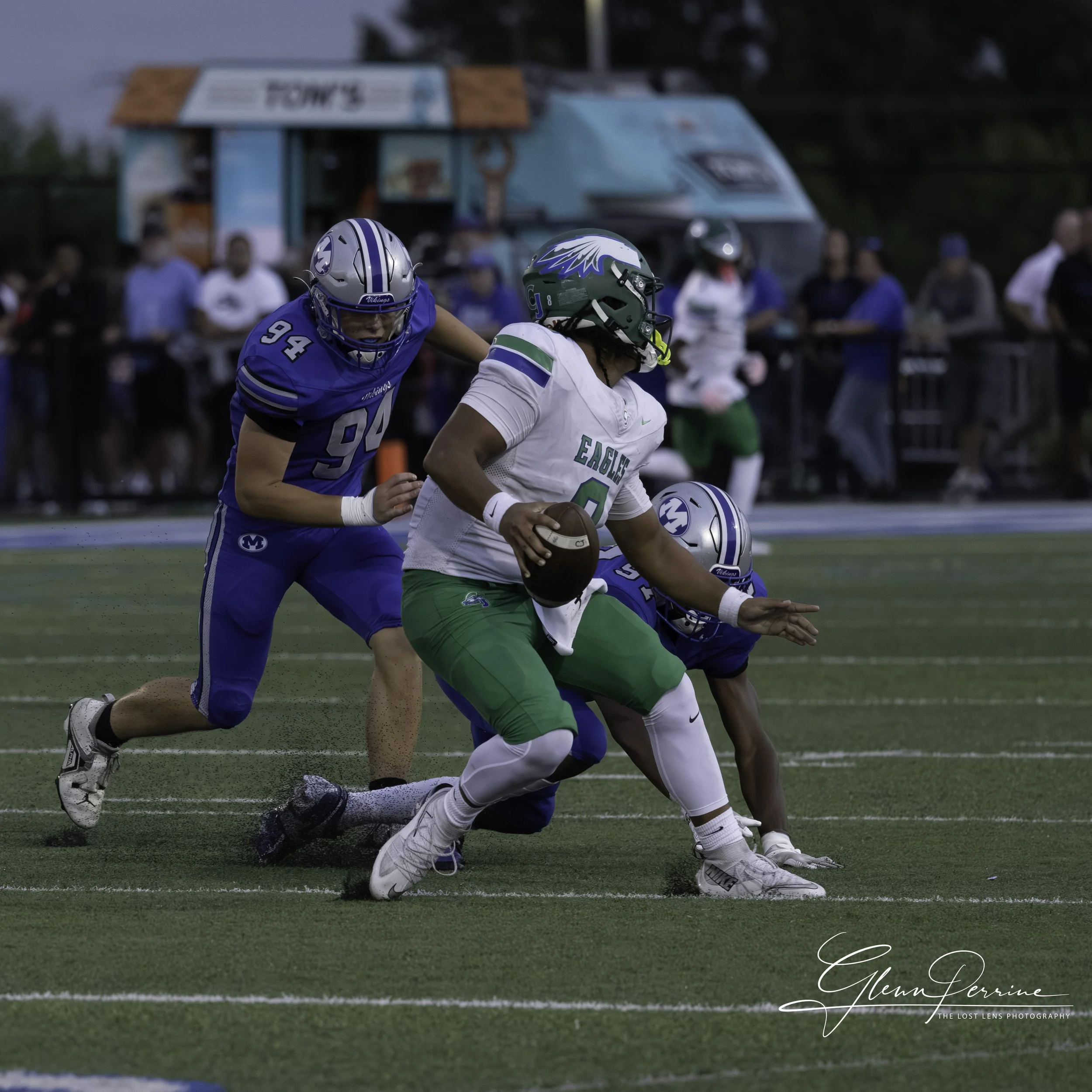 An American football game in progress with players from two teams: one in green and white uniform holding the football, and two in blue uniforms attempting to tackle the player. The players are on a green field with spectators in the background.