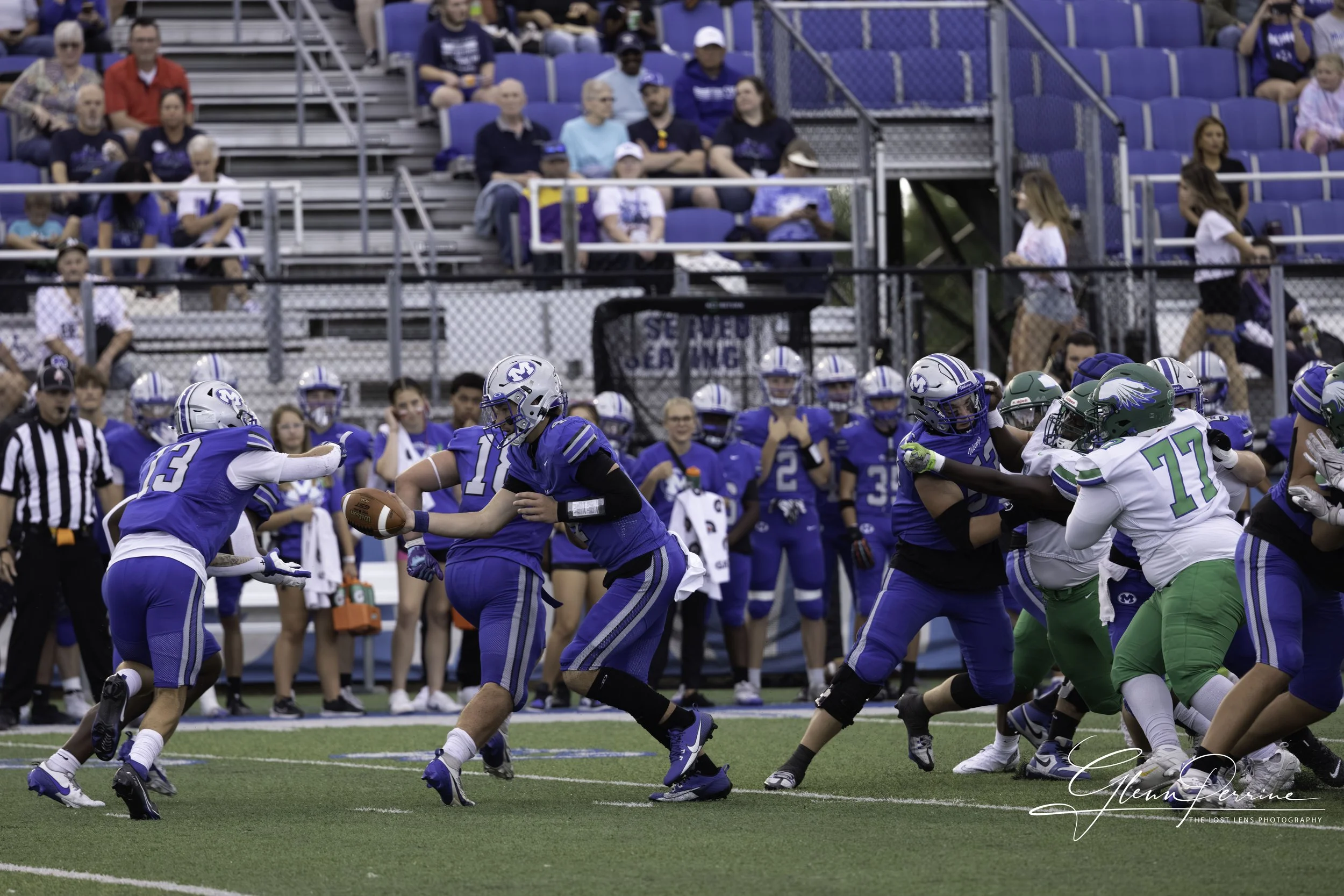 Football game with players in blue and green jerseys on the field, with spectators watching from the stands in the background.