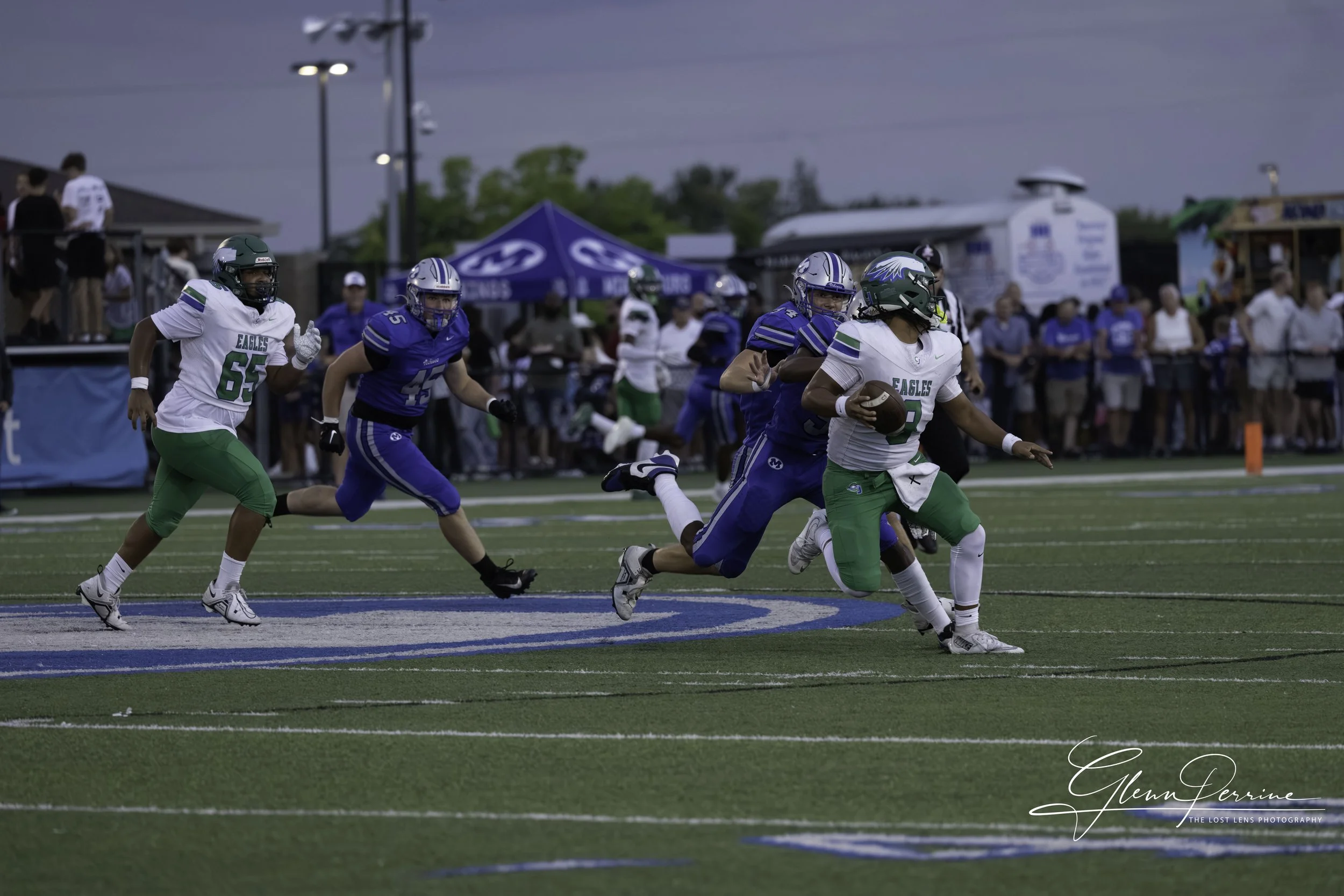 A high school football game with players in blue and green uniforms. The green team player with the ball is running towards the end zone, while players in blue attempt to catch up. Spectators are visible on the sidelines and in the background.