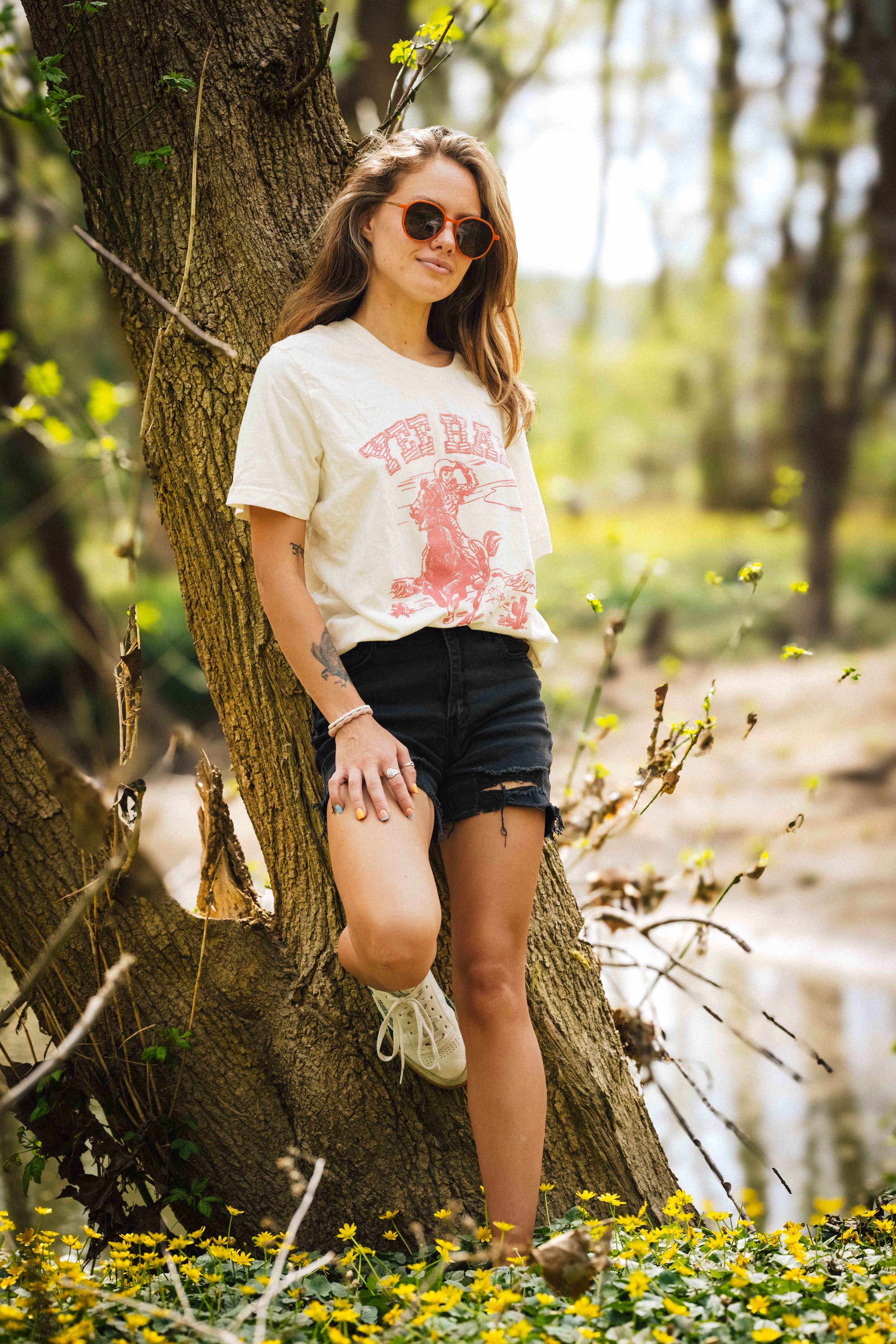 A young woman with long hair, wearing sunglasses, a vintage graphic T-shirt, black shorts, and white sneakers, leaning against a tree in a natural outdoor setting with yellow flowers in the foreground.