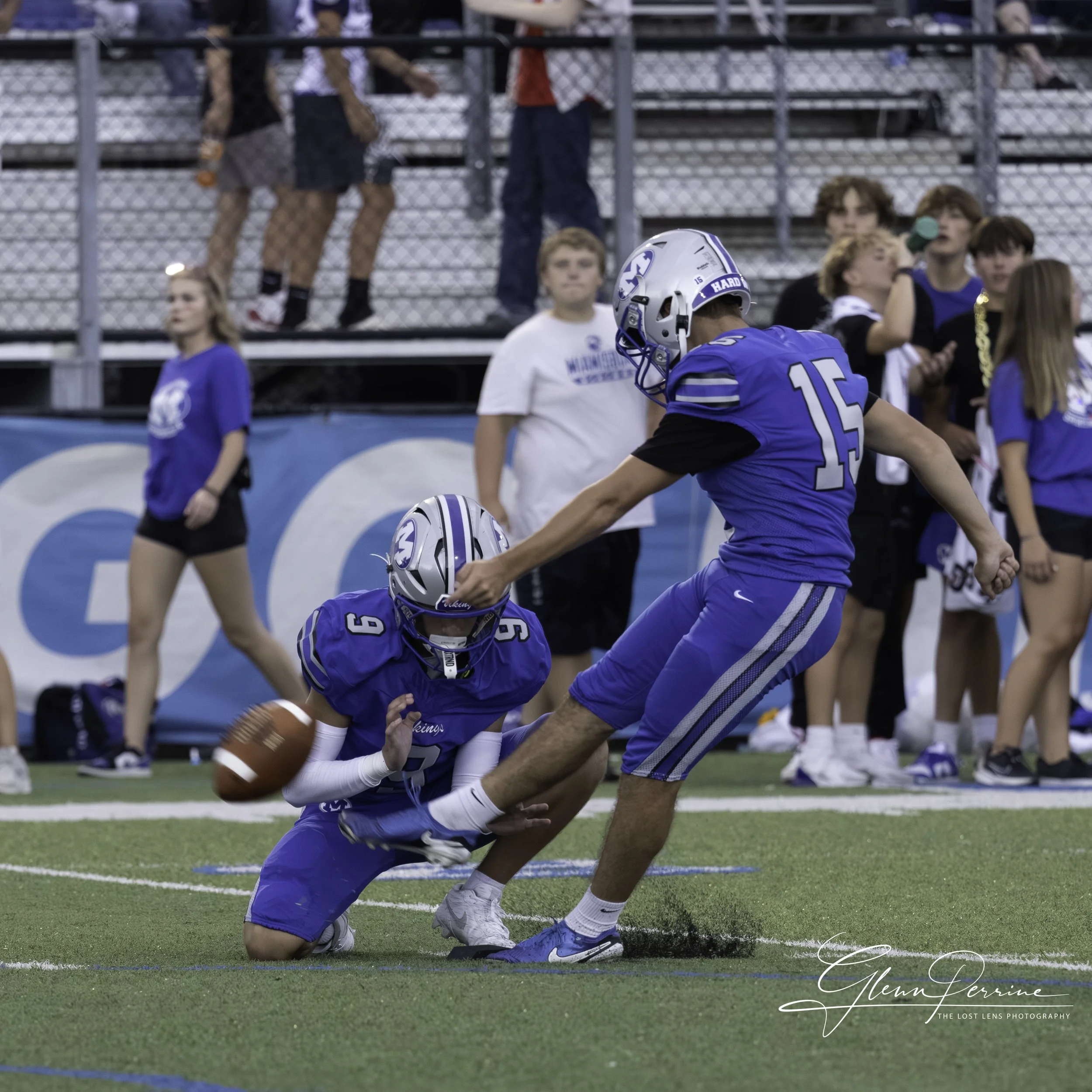 Football players in blue jerseys practicing a tackle on the field during a game, with spectators watching in the background.