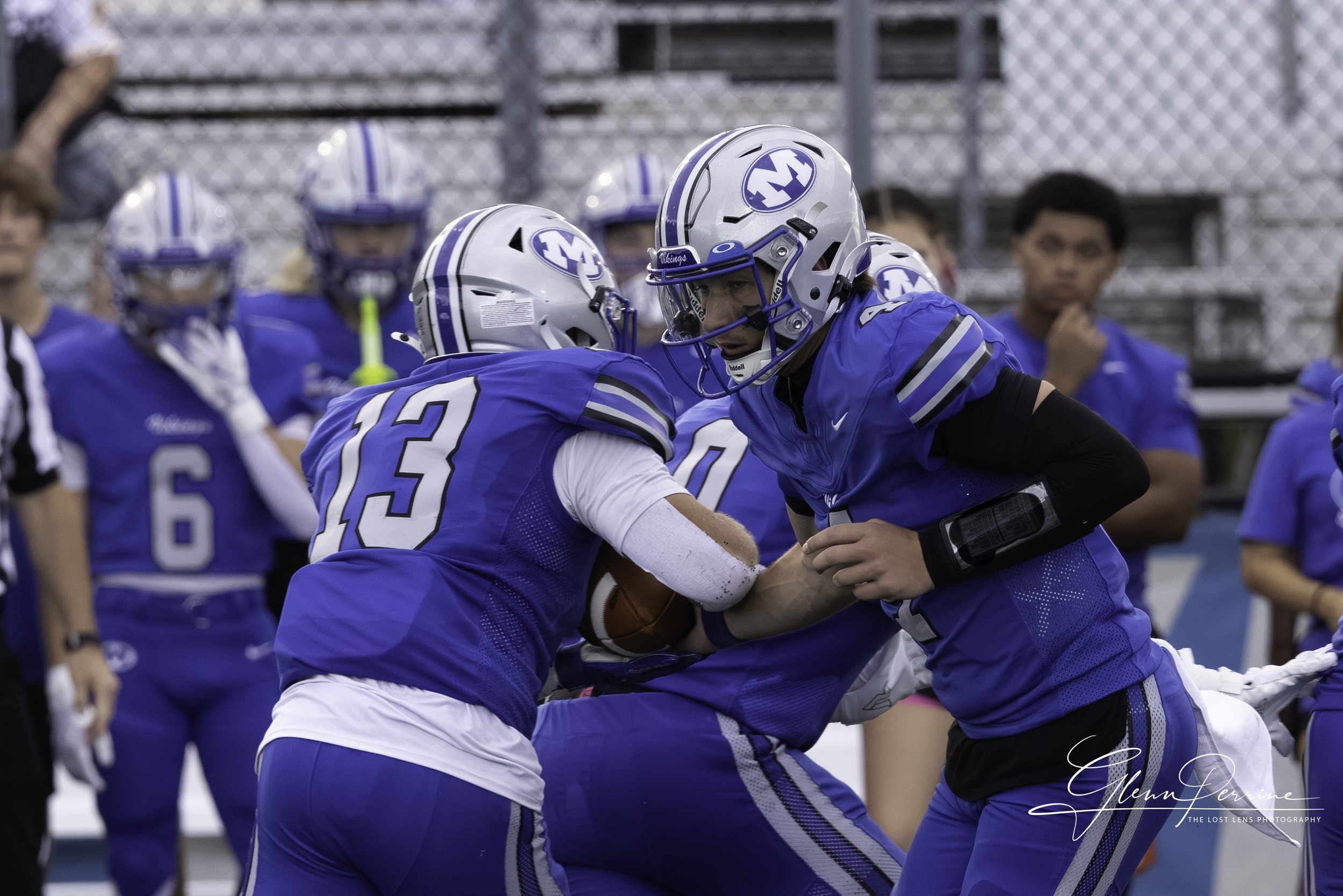Two football players in blue uniforms with helmets are engaging in a play during a game, with one player holding the football and the other preparing to block.