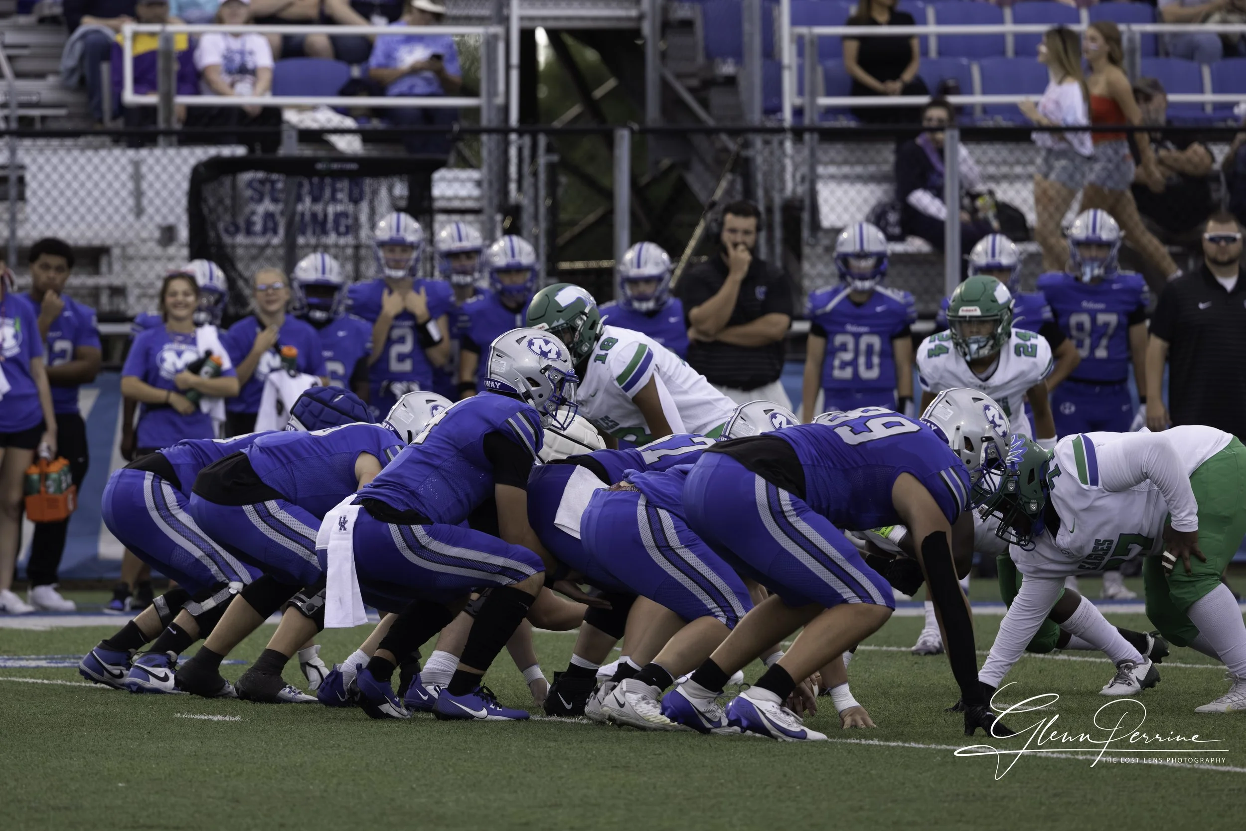 A youth football game on the field with players in blue and white uniforms, with spectators and team members watching from the bleachers.