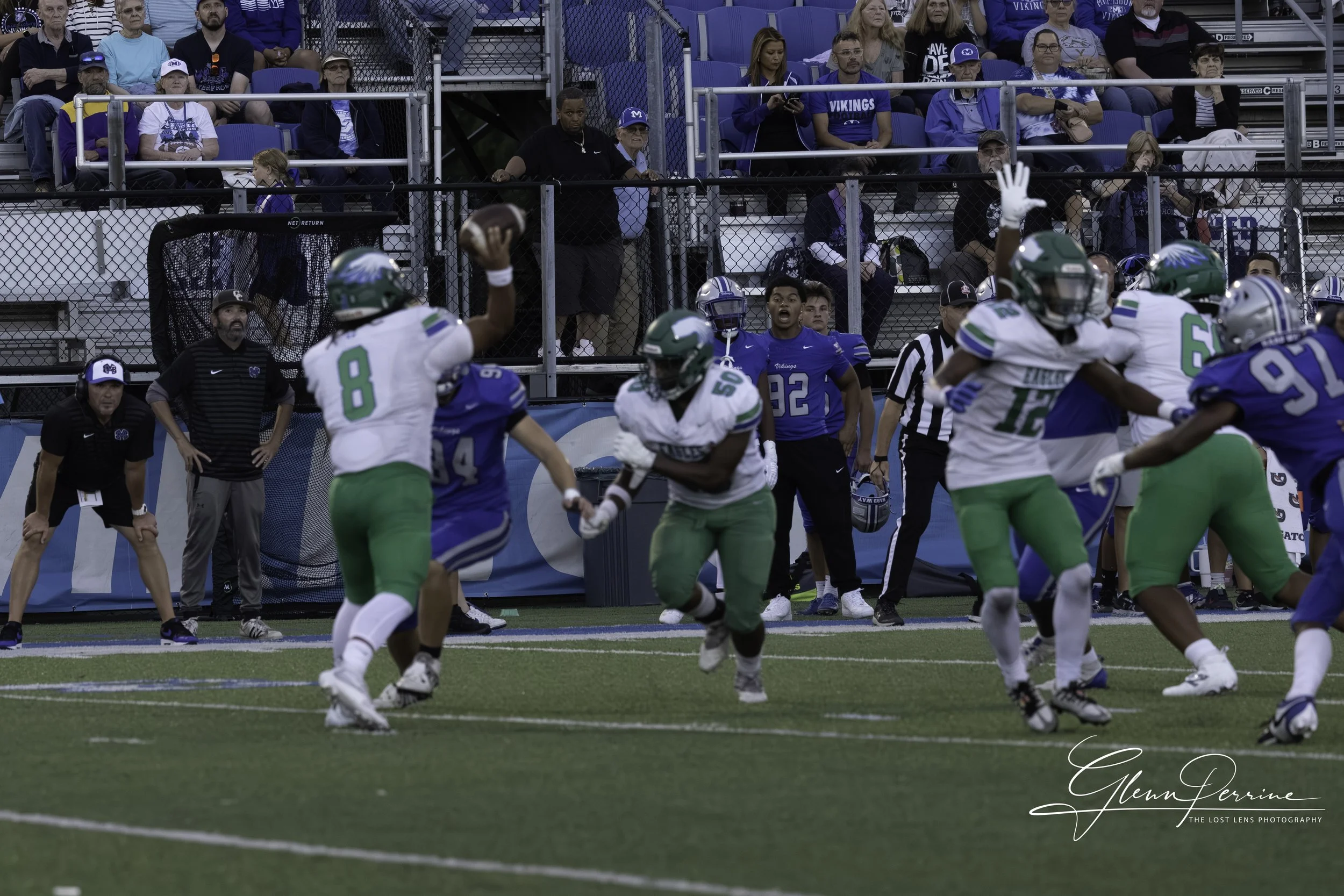 An American football game in progress with players from two teams on the field. One player in a white jersey with green pants is running with the ball, while others from the same team are blocking. Opponents in blue jerseys are attempting to stop the