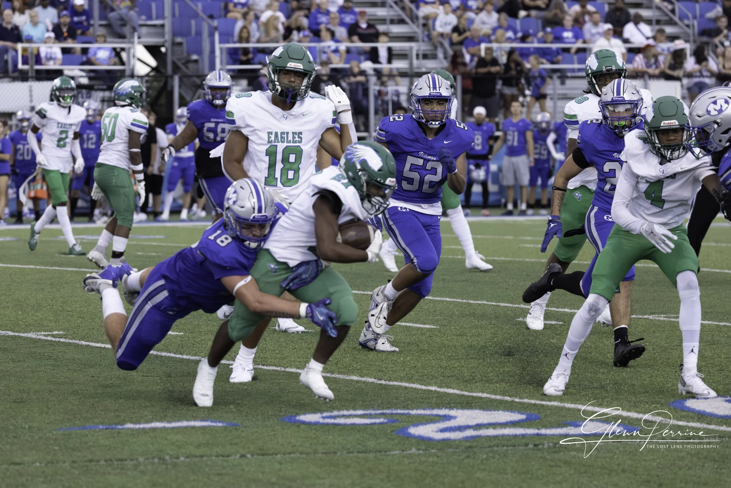A football game with players in blue and white uniforms on the field; one player in white is carrying the ball while a player in blue attempts to tackle him, with spectators watching from the stands in the background.
