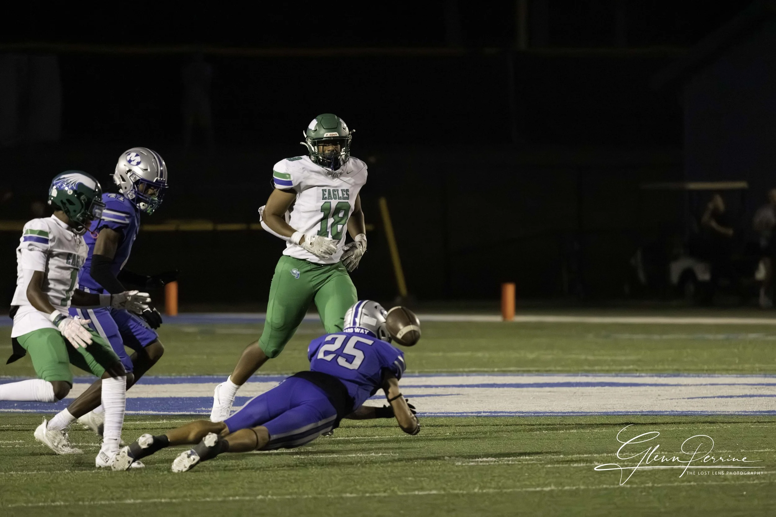 Football players during a game on a night field. One player in a green and white uniform with the number 18 stands while another in a blue uniform with the number 25 is on the ground, reaching for the football. Two other players are nearby, one in wh